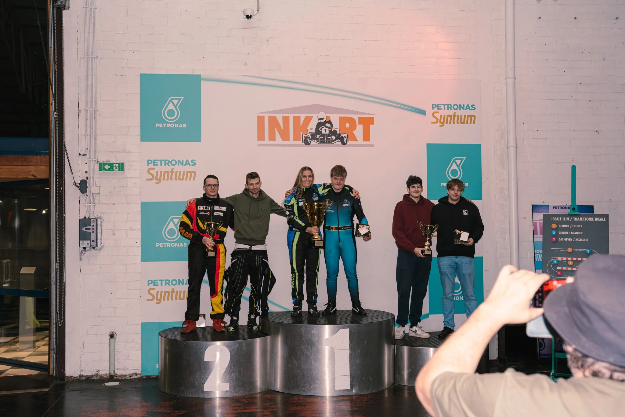 Six individuals on a winners' podium holding trophies at a go-kart racing event, with a backdrop displaying the event logo and sponsors.