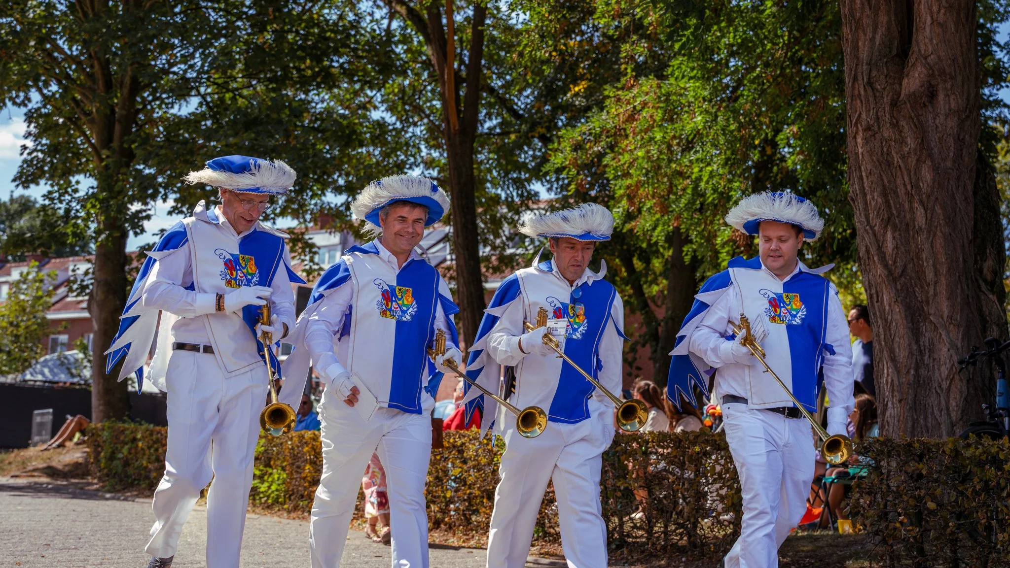 Four men dressed in blue and white uniforms with large feathered hats, carrying brass instruments, walking outdoors during a parade or celebration.