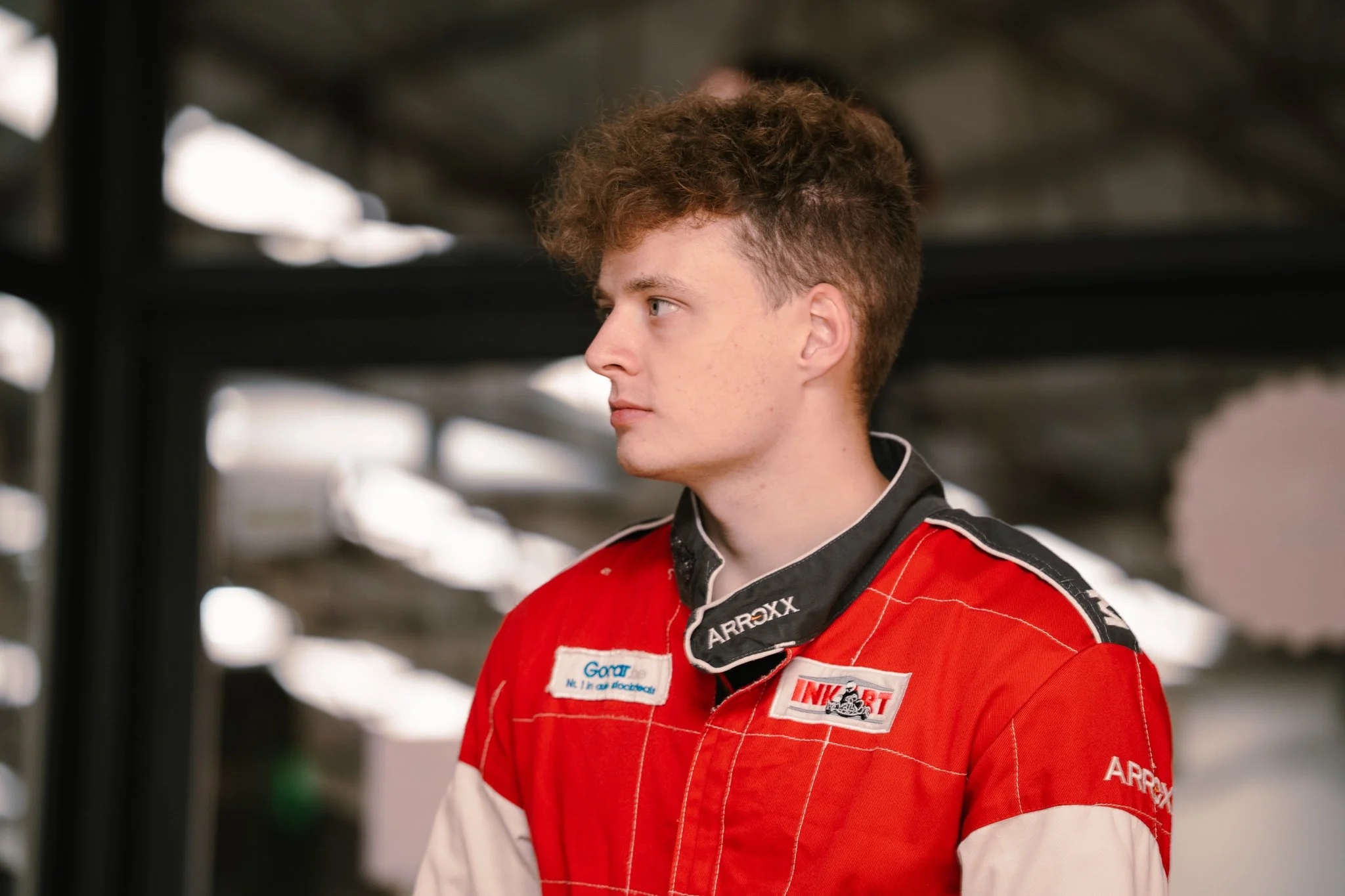 Young race car driver in red racing suit and black neck cover in garage.