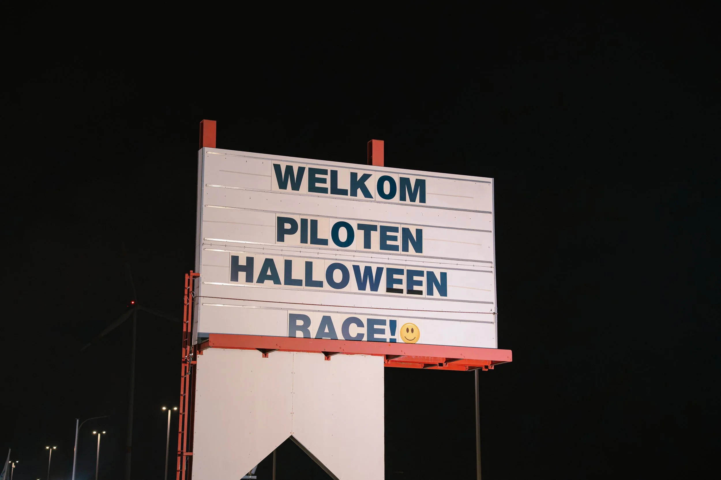 A large signboard with the words 'Welkom Piloten Halloween Race' displayed, with a smiley face emoji at the bottom, set against a dark night sky.
