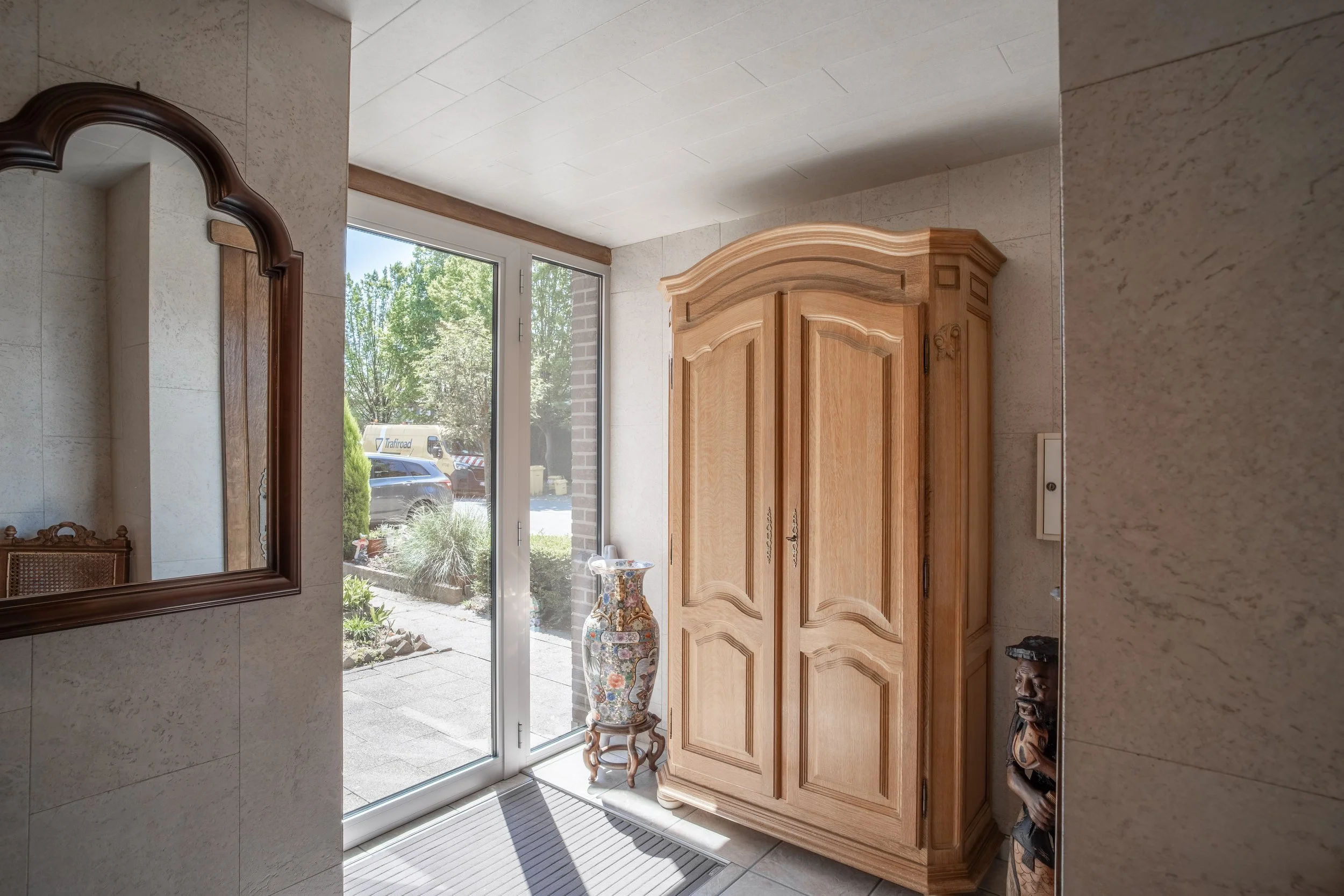 A view from inside a house showing a wooden cabinet, a large decorative vase, a wall mirror, and a sliding glass door leading outside, with trees and parked vehicles visible through the window.