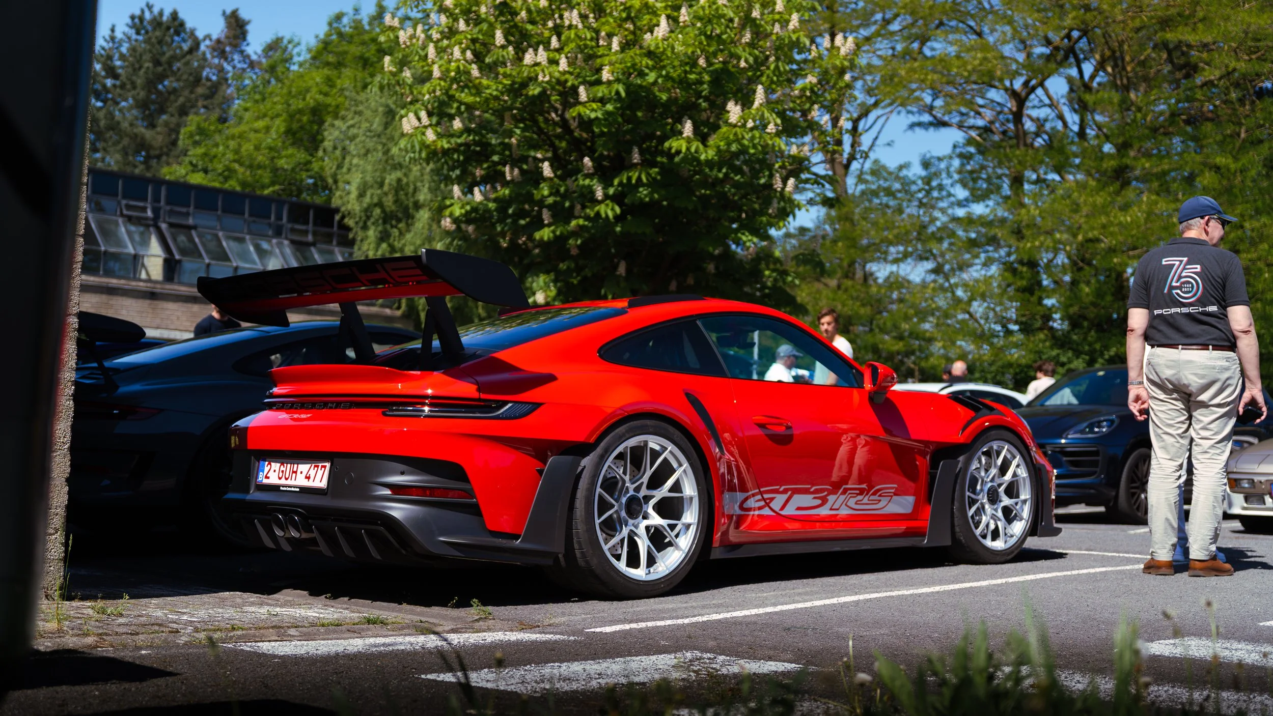 Red Porsche 911 GT3 RS parked among other cars at a car meet, with a man in a Porsche 75th anniversary shirt observing nearby, green trees and a building in the background.