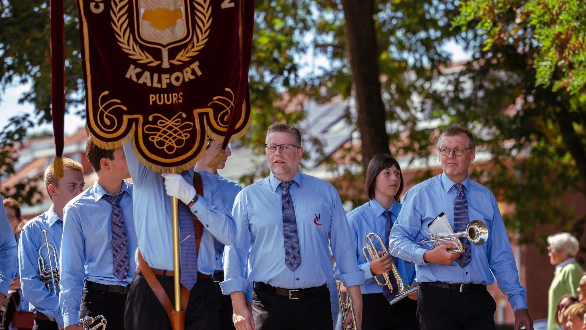 A group of people dressed in light blue shirts and dark pants, participating in a parade. One person is holding a large maroon banner with gold embroidery and the words 'KALFORT PUURS.' Many carry brass instruments. Trees and buildings are visible in