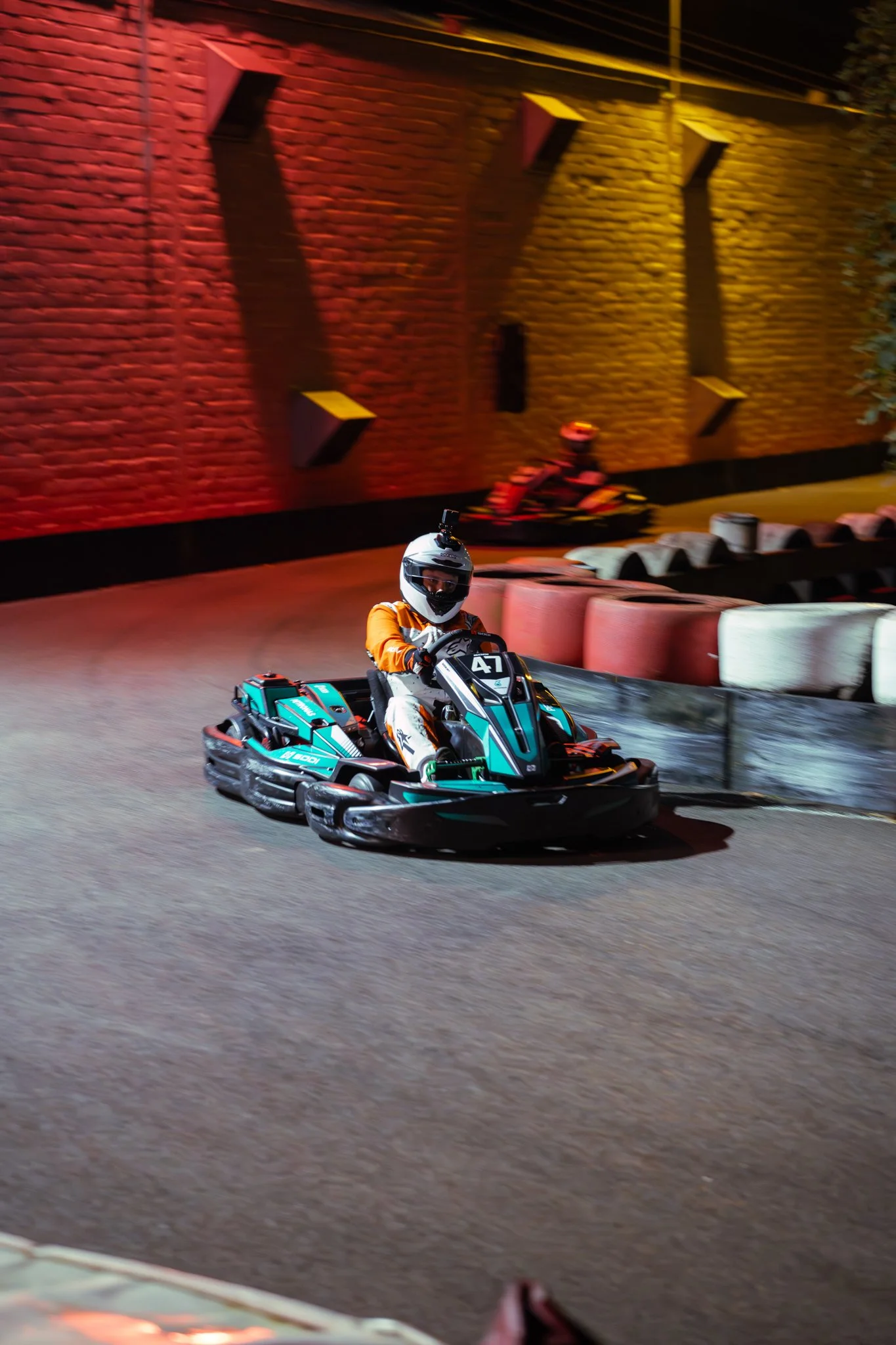 A person in racing gear and helmet driving a go-kart on an indoor track at night, with a brick wall and another kart in the background.
