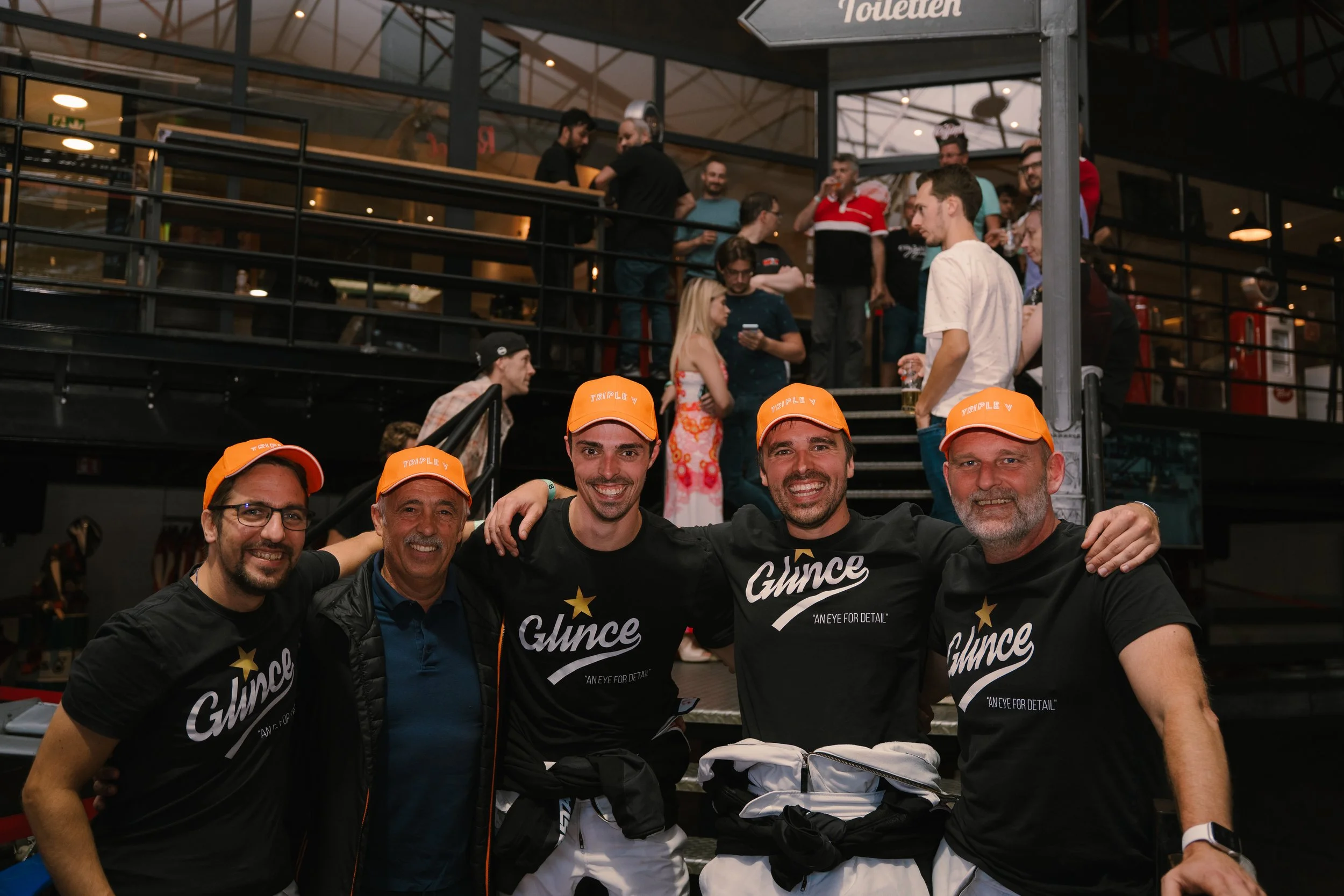 A group of five men smiling and wearing black T-shirts with 'Glyce' and 'Aye For Detail' printed on them. They are wearing orange caps saying 'TRIPLE V' and are posing in front of a crowd of people at an indoor event.