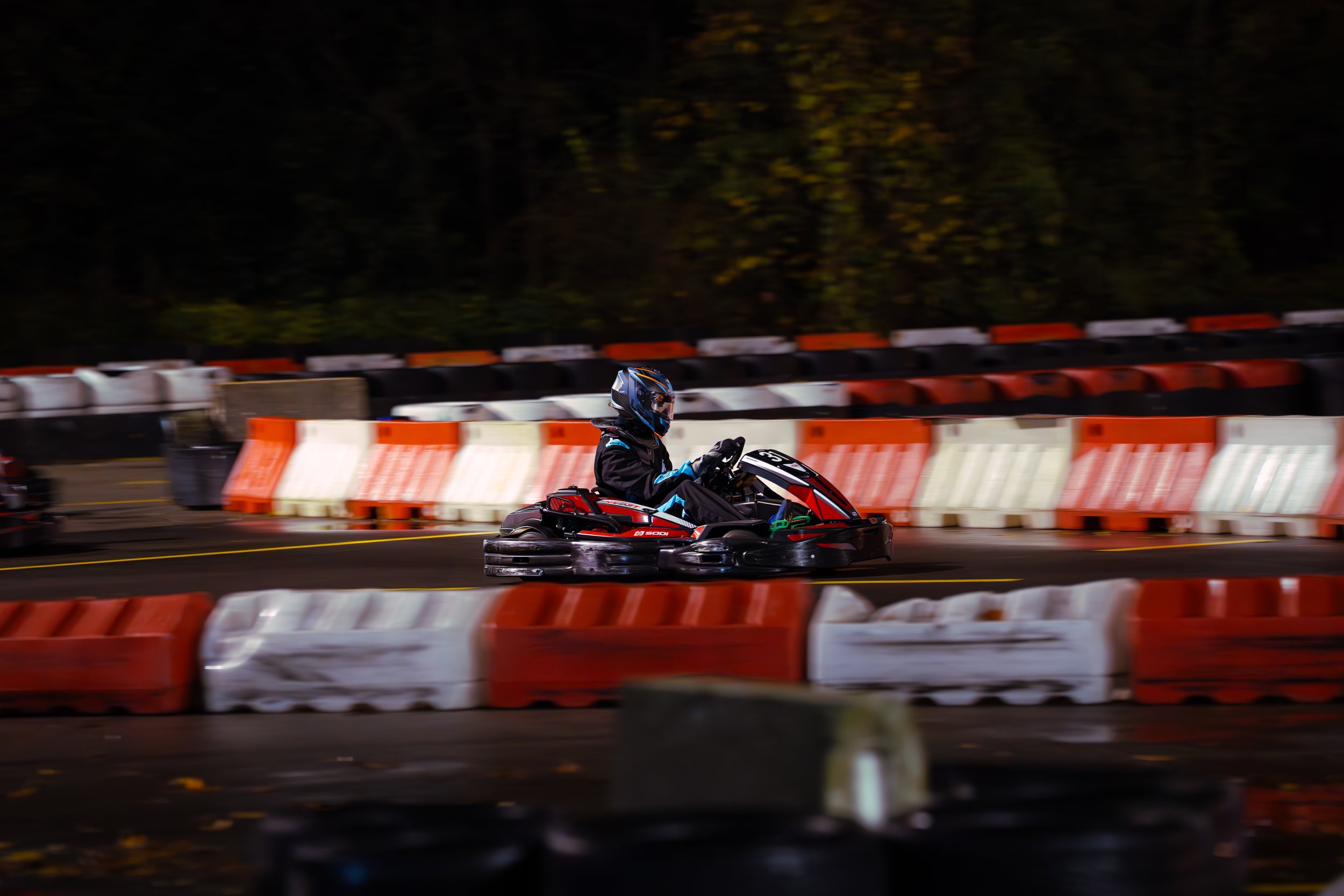 A person wearing a helmet and riding a go-kart on an outdoor track at night, with red and white barriers surrounding the track.