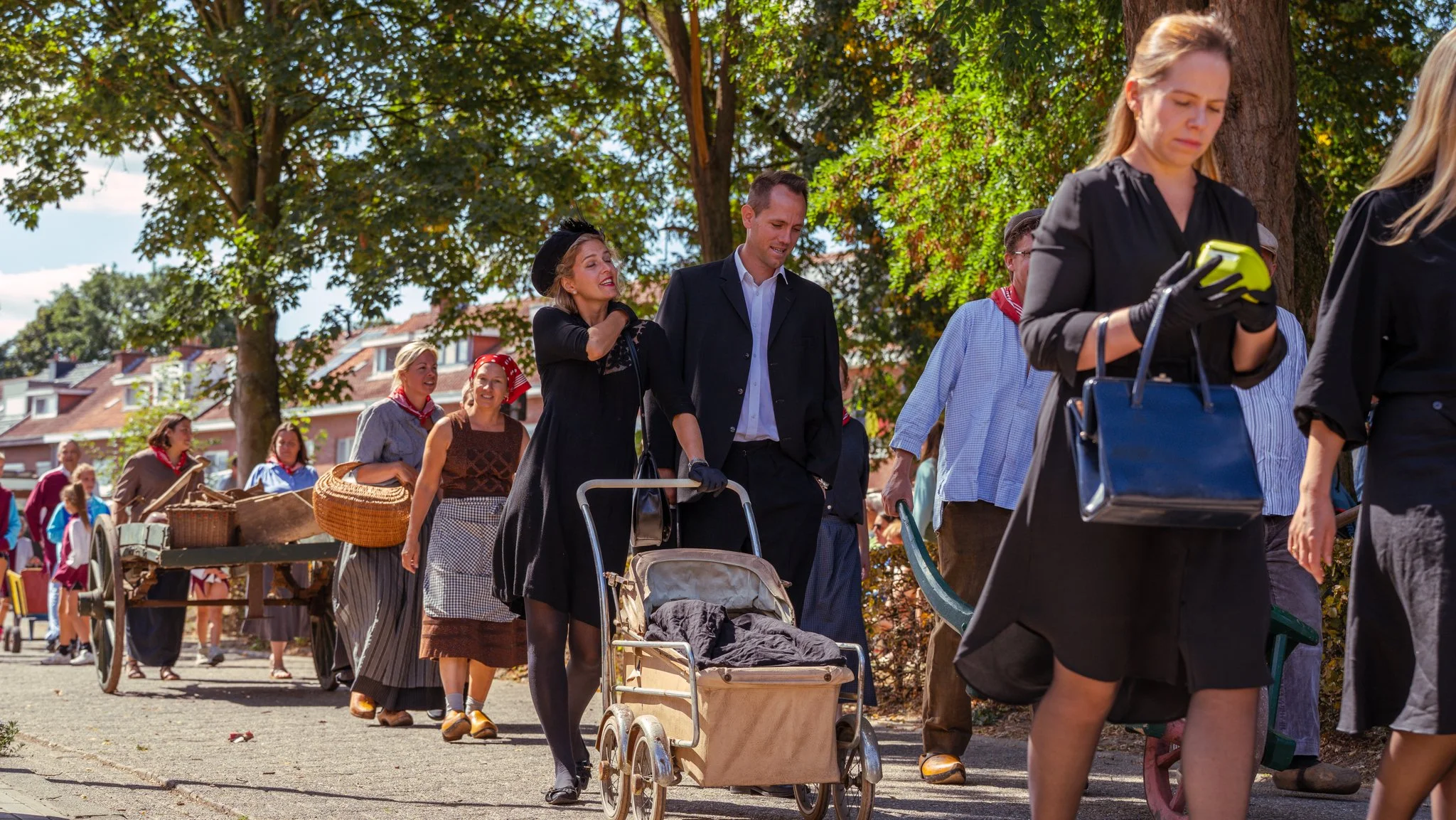 People dressed in historical clothing participate in a parade or festival outdoors amid trees and houses.