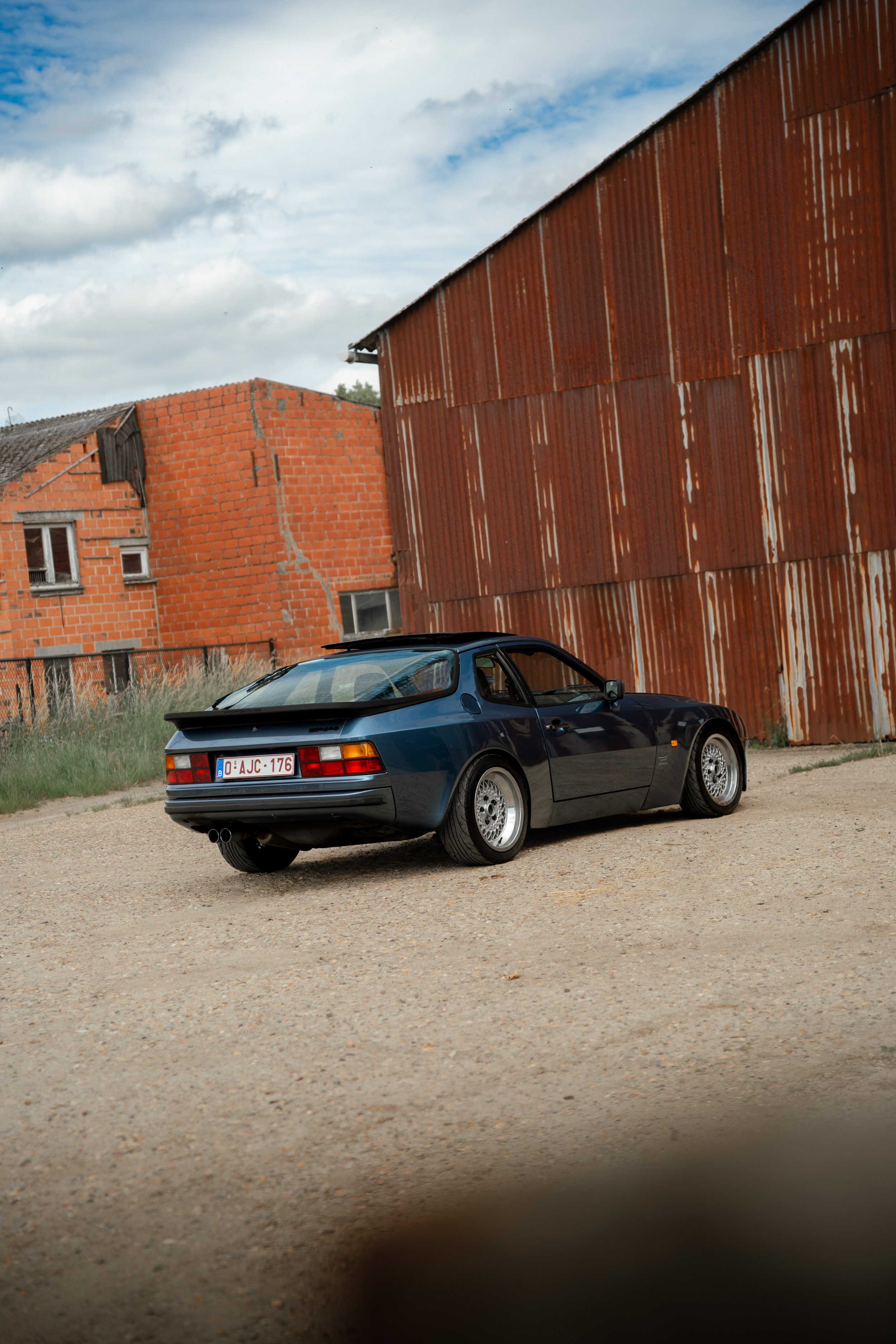 A dark blue vintage sports car parked on a gravel lot in front of an unfinished brick building and a rusty metal shed under a partly cloudy sky.