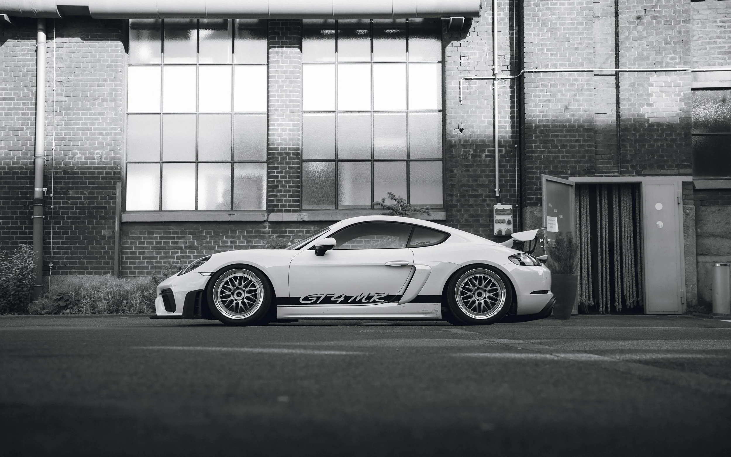 A white sports car with black and white racing decals parked in front of an industrial brick building with large industrial windows and pipes.