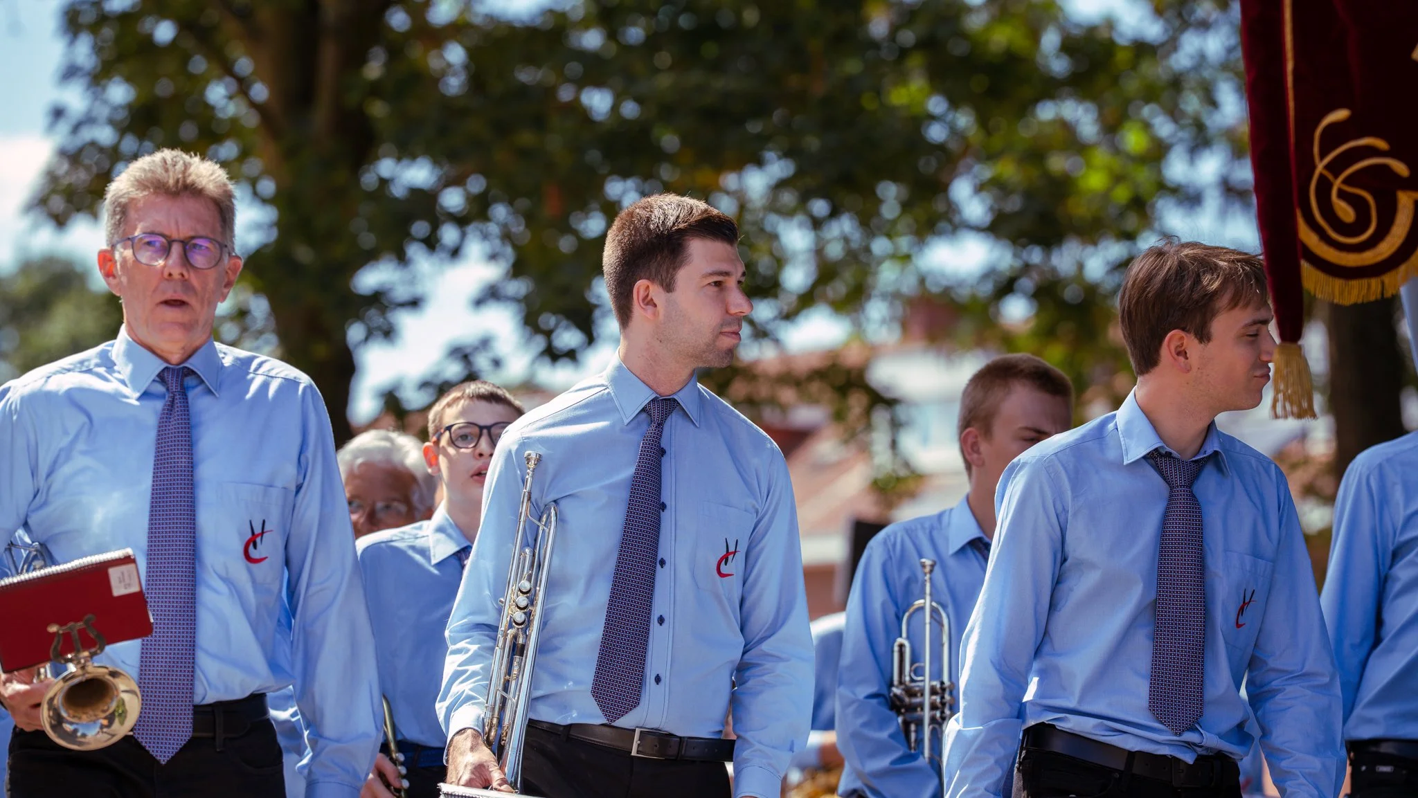 A group of people in blue shirts and ties, some holding musical instruments, standing outdoors during a parade or procession.