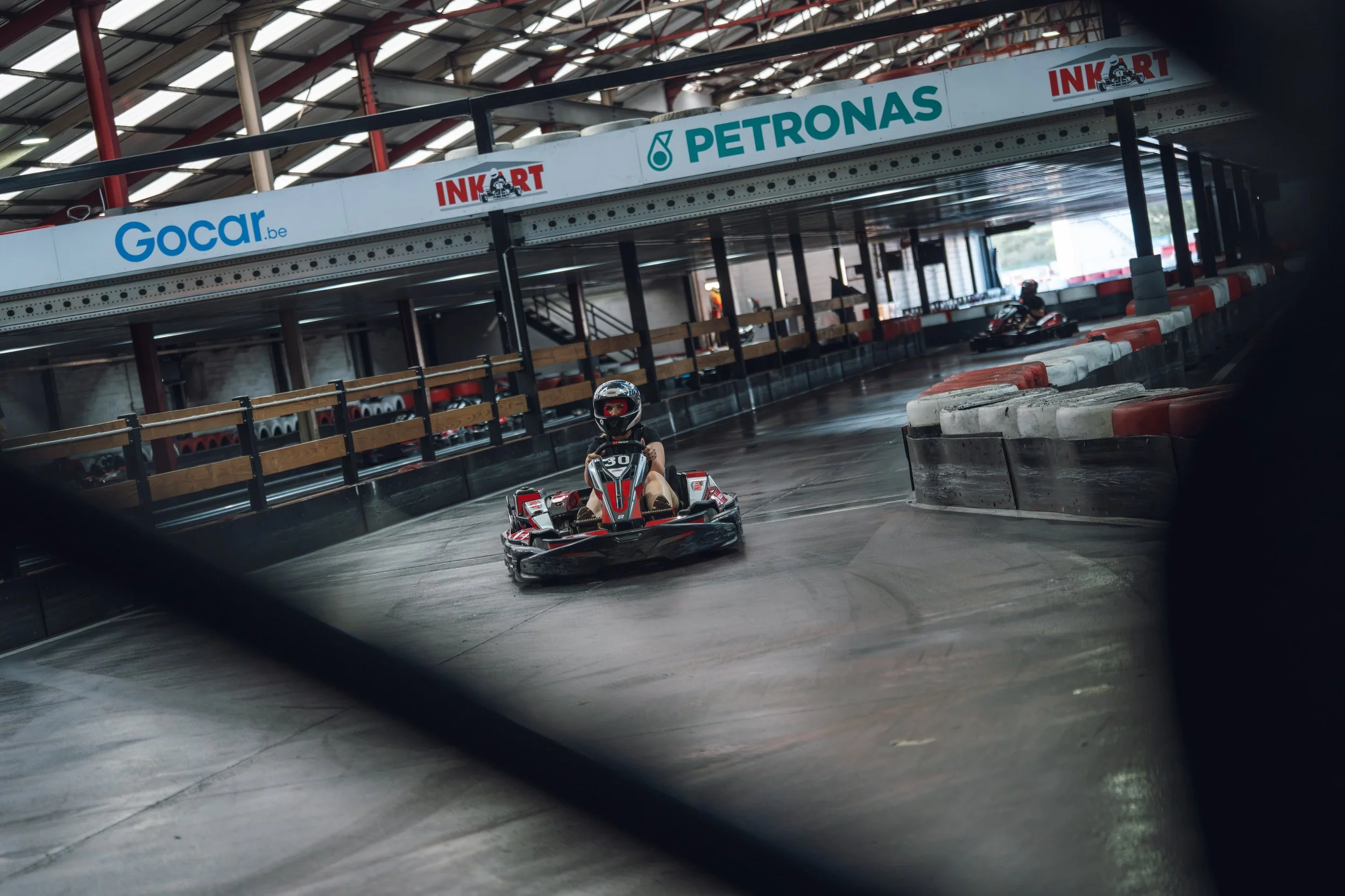Indoor go-kart track with a driver in a black helmet and red go-kart, barriers lining the track, and sponsorship banners overhead including Gocar.be and Petronas.