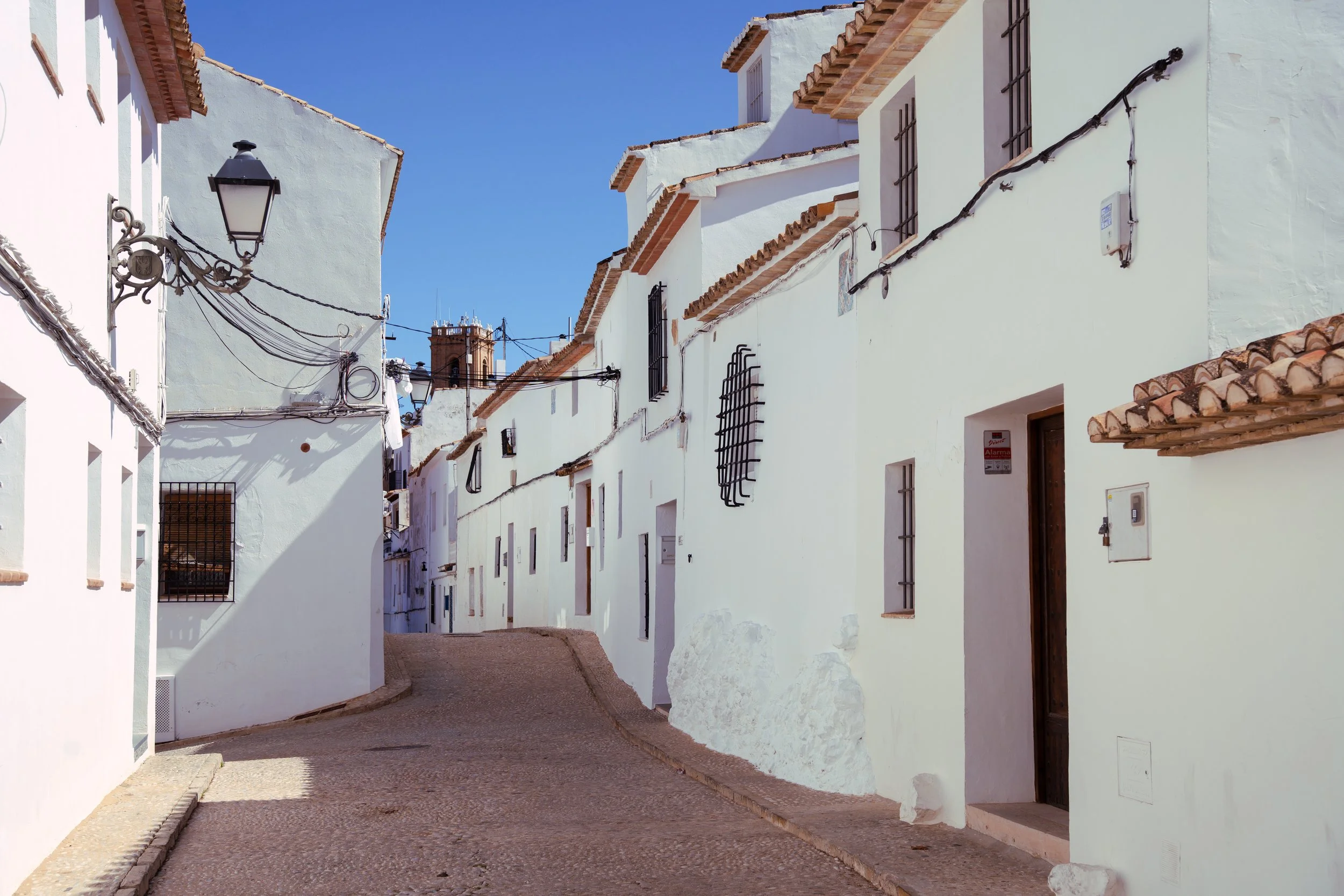 A narrow, winding street in a white Mediterranean village with terracotta-tiled roofs and iron window grilles, under a bright blue sky.