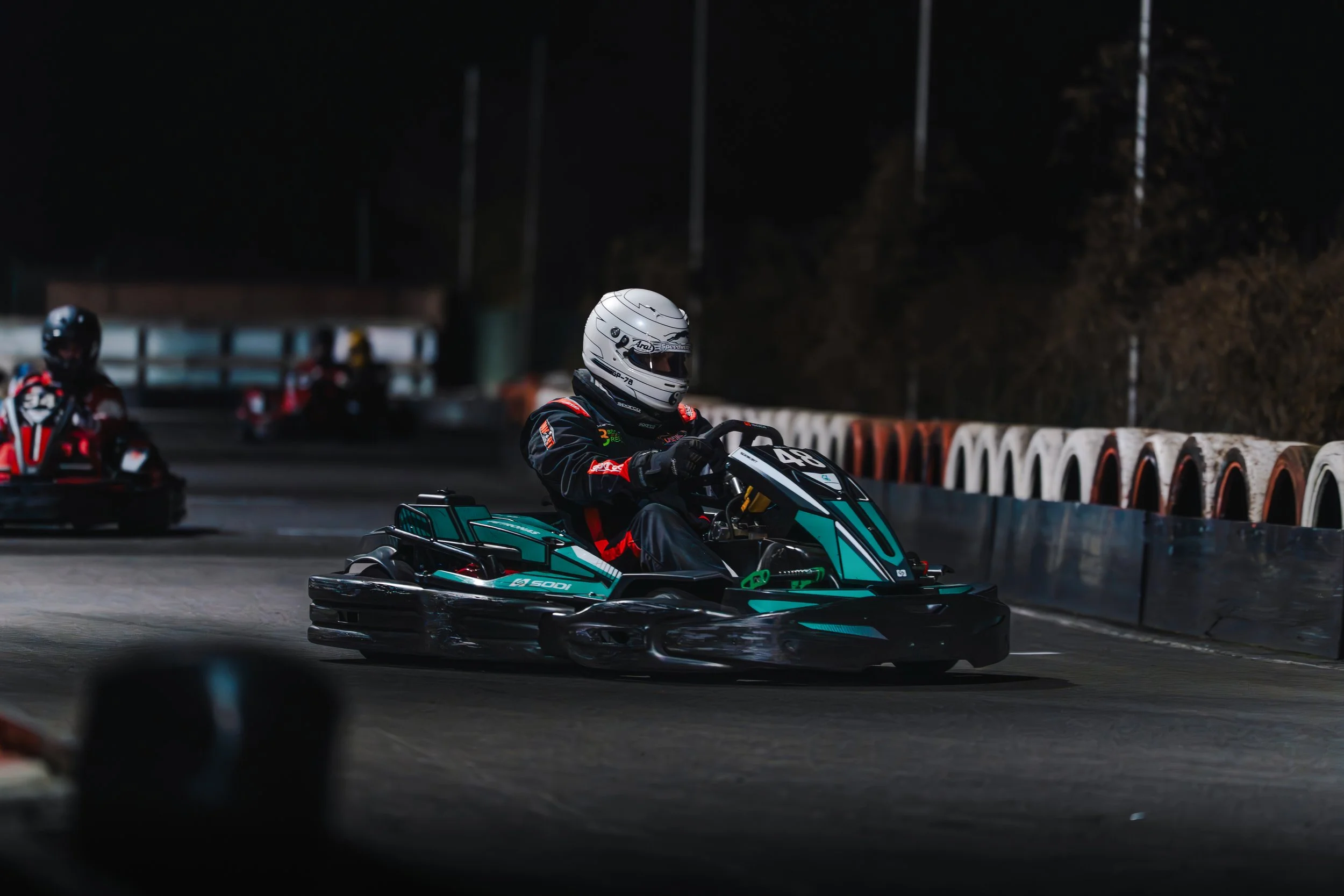 Race car drivers in racing suits and helmets driving go-karts on a track at night.