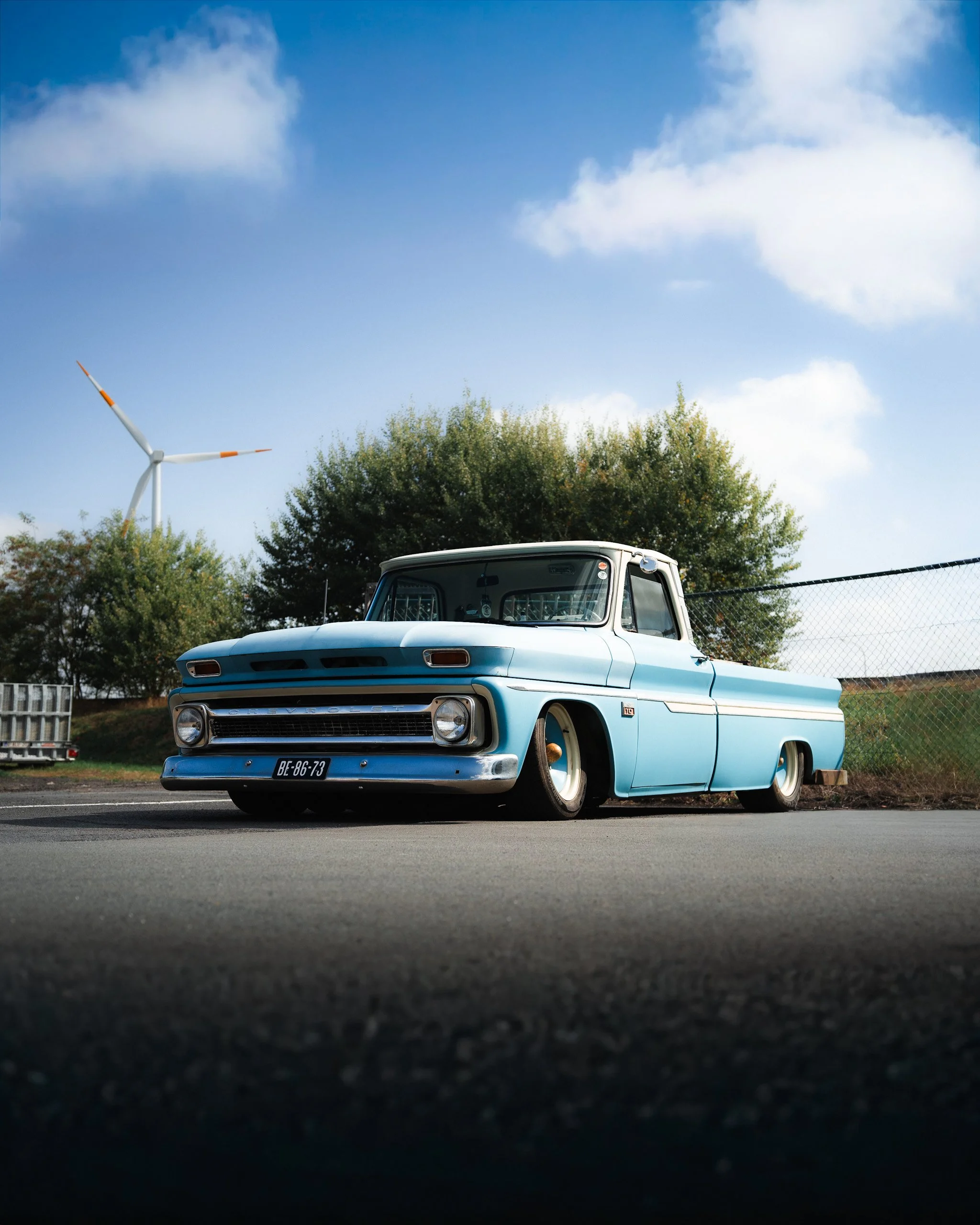 A vintage light blue pickup truck parked on the side of the road, with trees, a wire fence, and a wind turbine in the background under a partly cloudy sky.