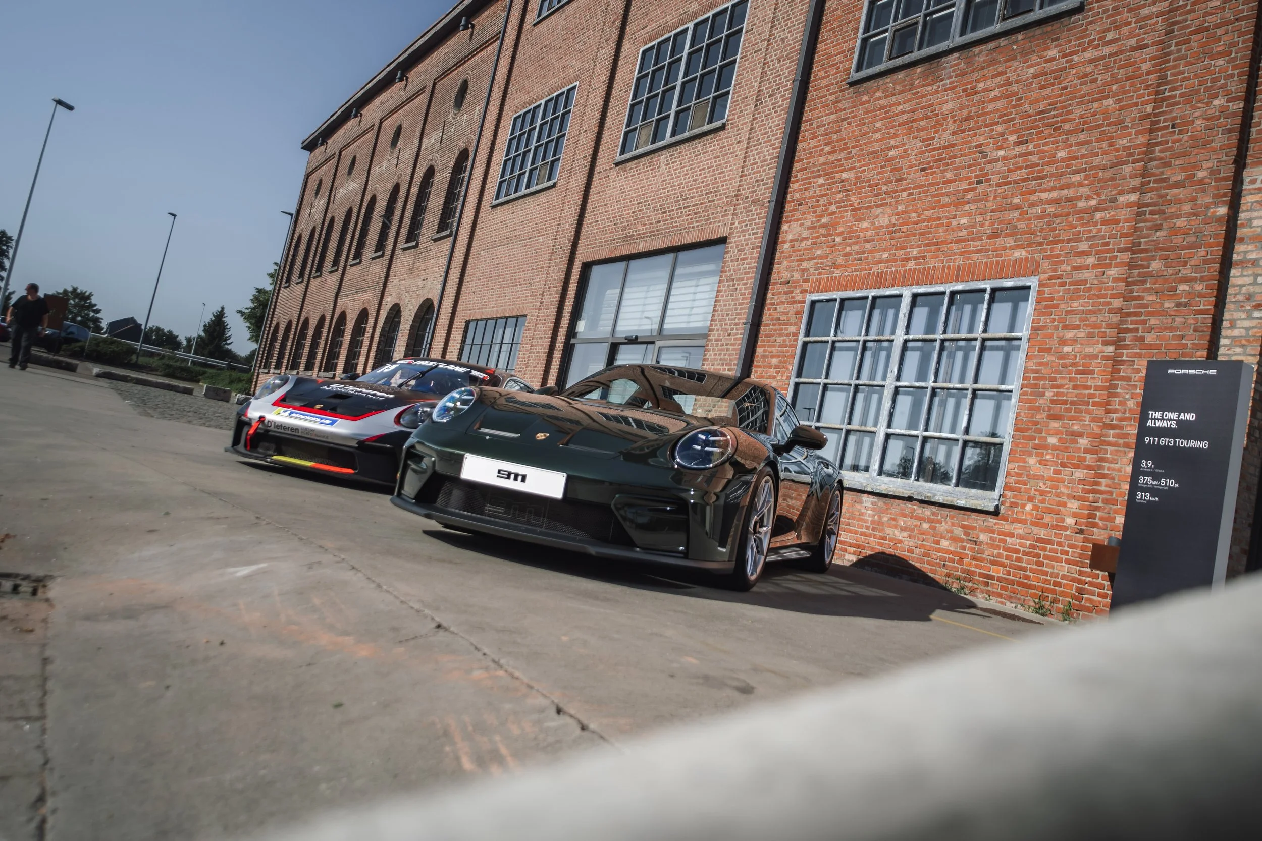 Two Porsche cars parked in front of a red brick industrial building with large windows, under a clear blue sky.