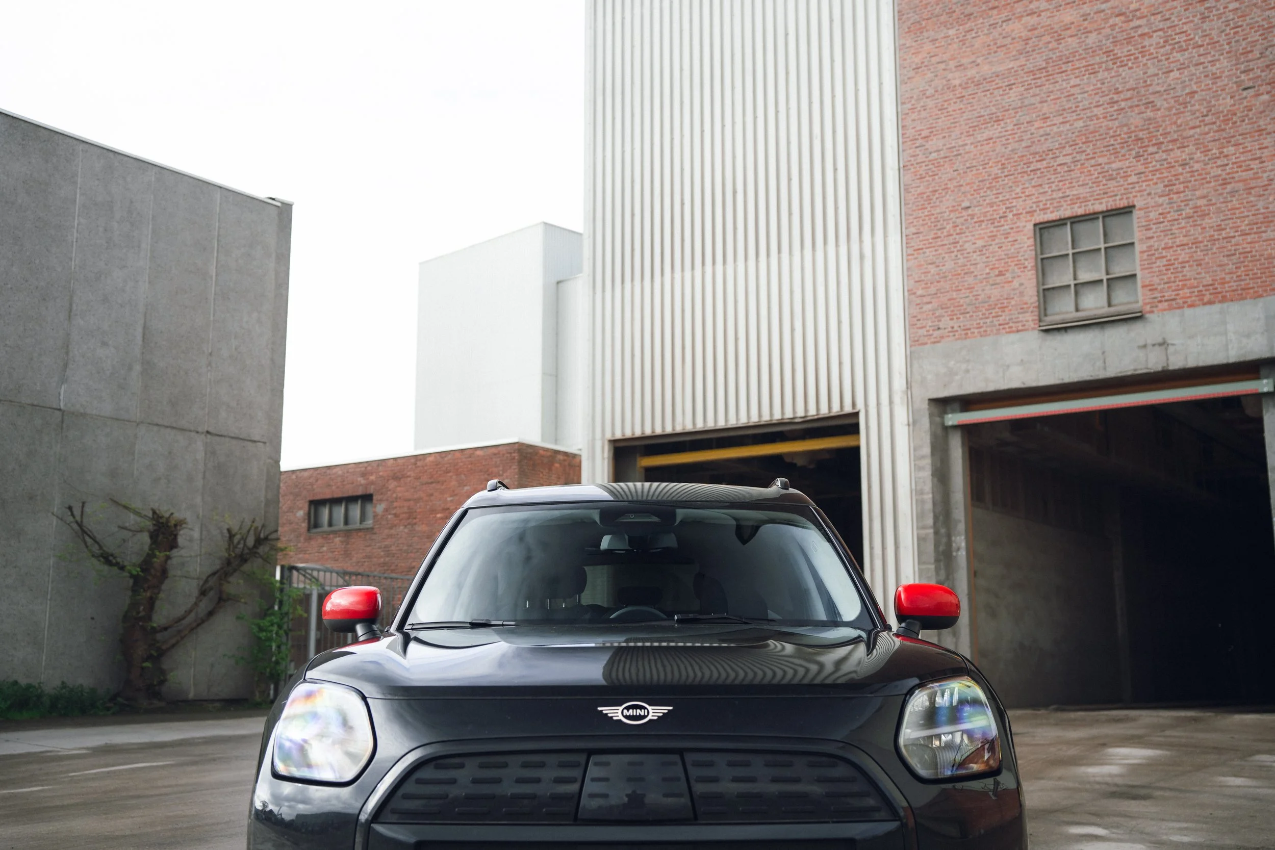 Black Mini Cooper with red side mirrors parked outside industrial buildings with brick, concrete, and metal siding.