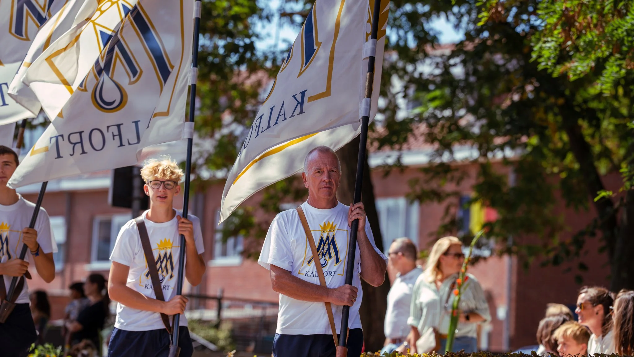 People participating in a flag procession outdoors, carrying white flags with a crest and the words 'Kalovrt' on them, with trees and buildings in the background.