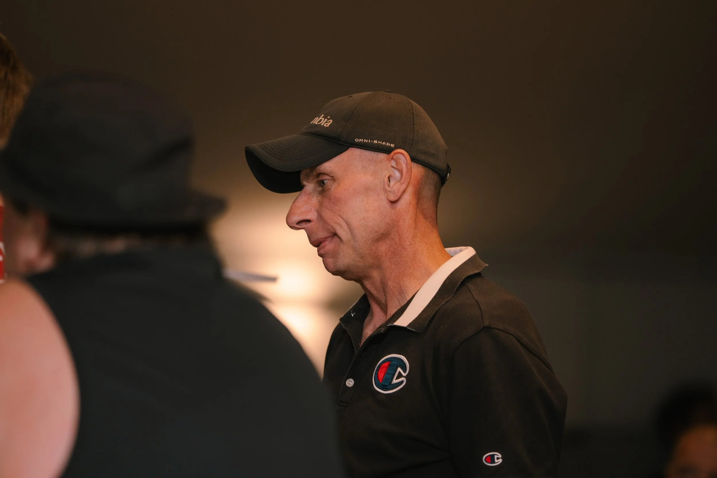 A man wearing a black cap and black shirt with a Champion logo is engaging in conversation with a person whose back is visible, wearing a black hat and black clothing, in an indoor setting with a dark background.