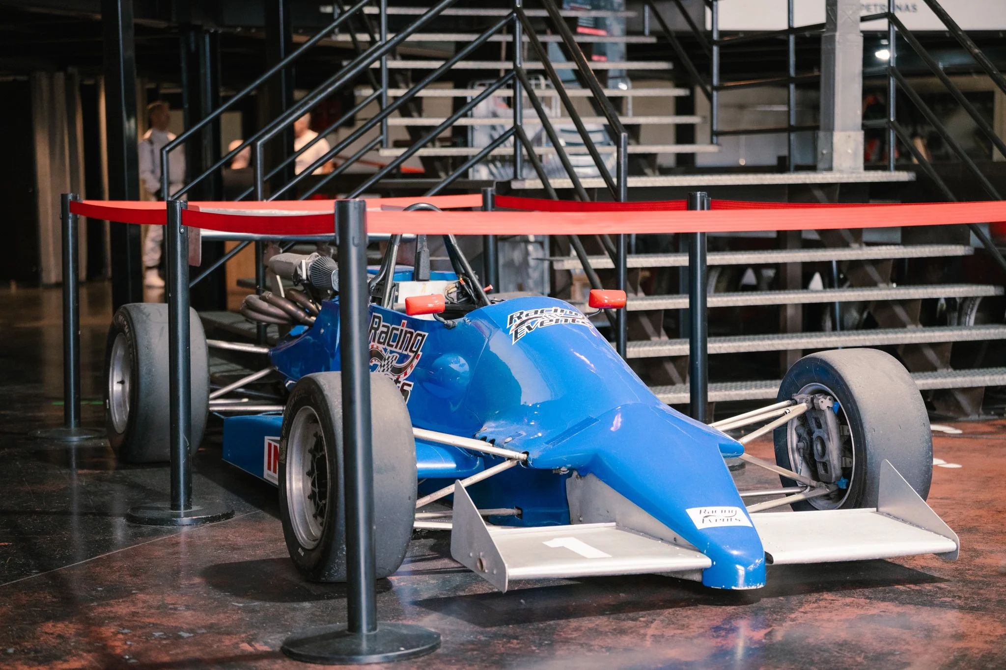 A vintage blue open-wheel race car displayed behind a red rope barrier at an indoor exhibit, with stairs and people in the background.