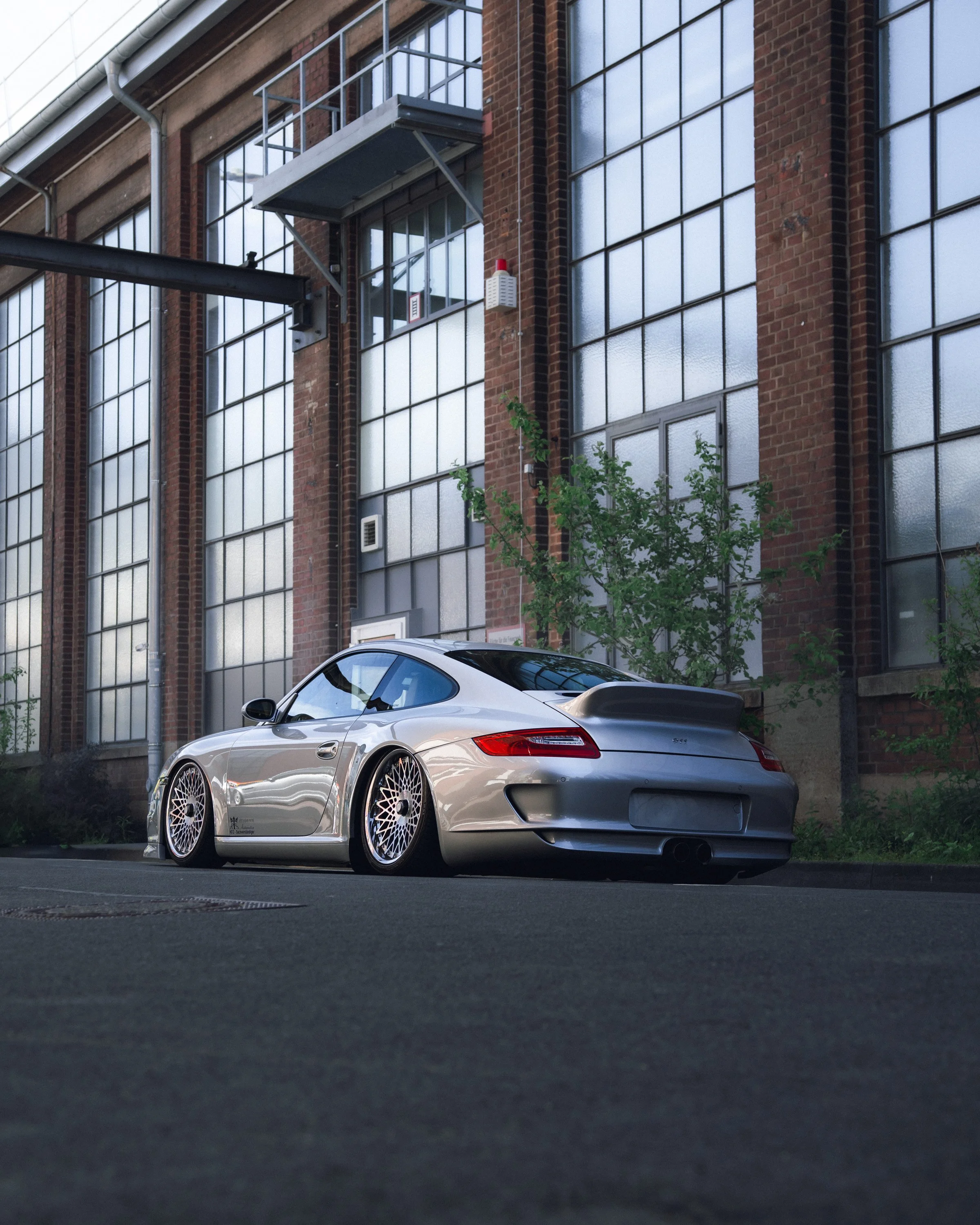 Silver sports car parked in front of a brick industrial building with large grid windows and a small tree in the foreground.