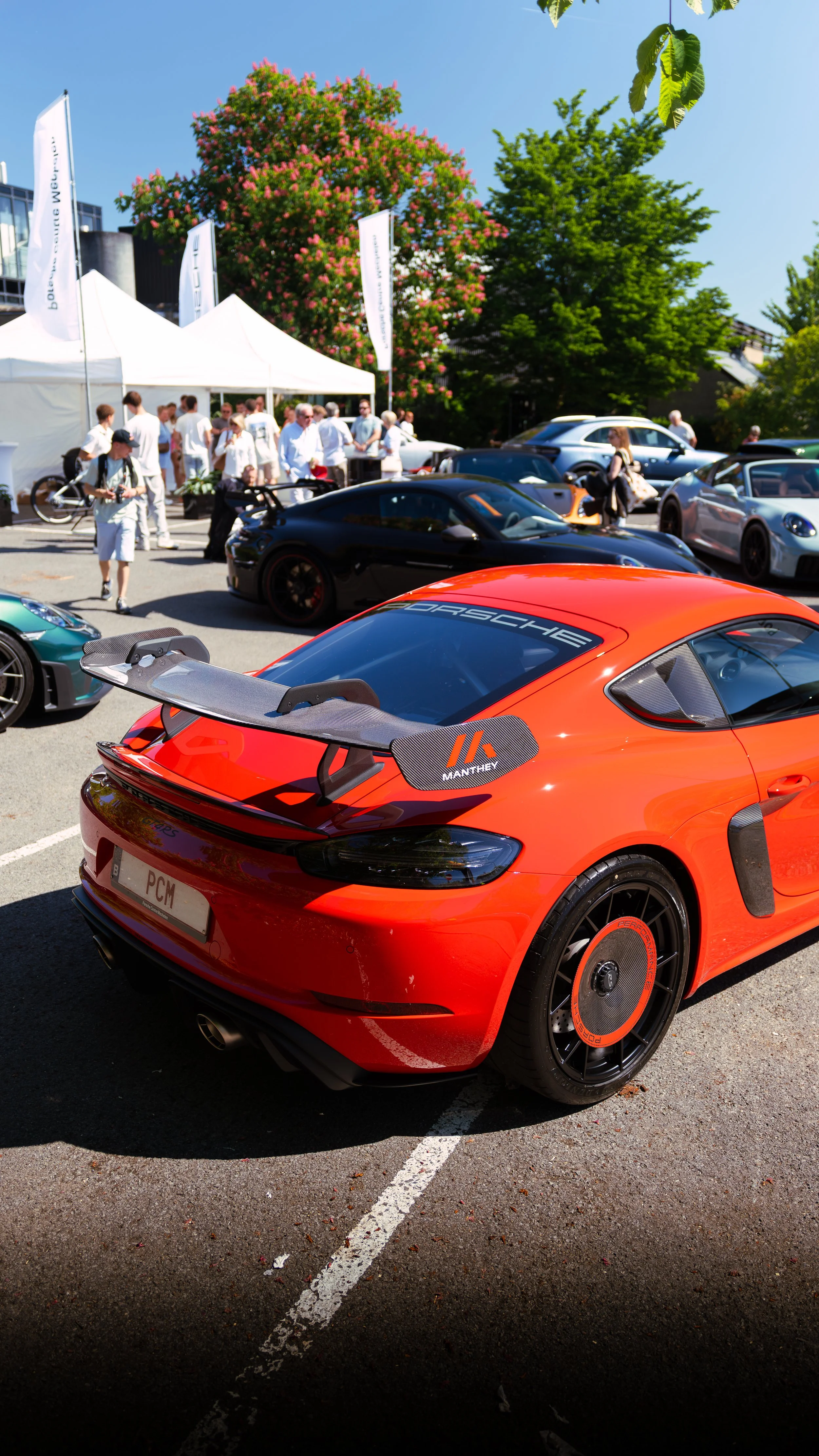 Red Porsche sports car with a large rear wing at an outdoor car show, surrounded by other cars and attendees, with tents and trees in the background.