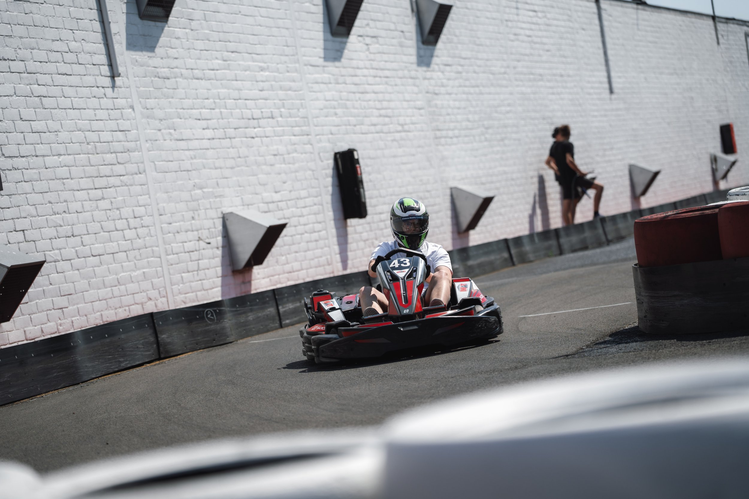 A person wearing a helmet and white shirt is driving a go-kart on a race track. The track has a white brick wall with black barriers and a person standing on the side, blurred in the background.