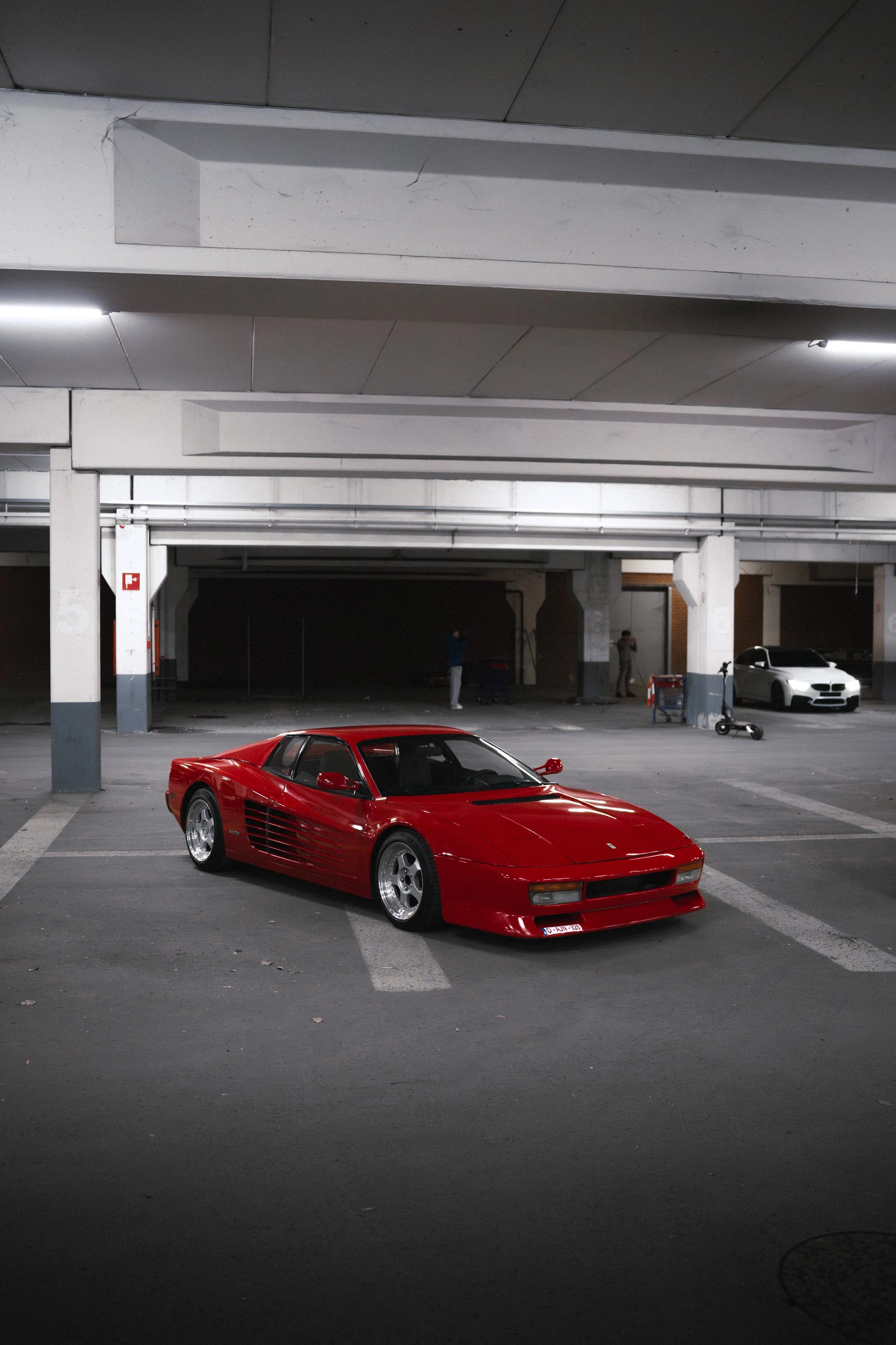 A red Ferrari sports car parked in an indoor parking garage, with white concrete pillars and a dark ceiling.