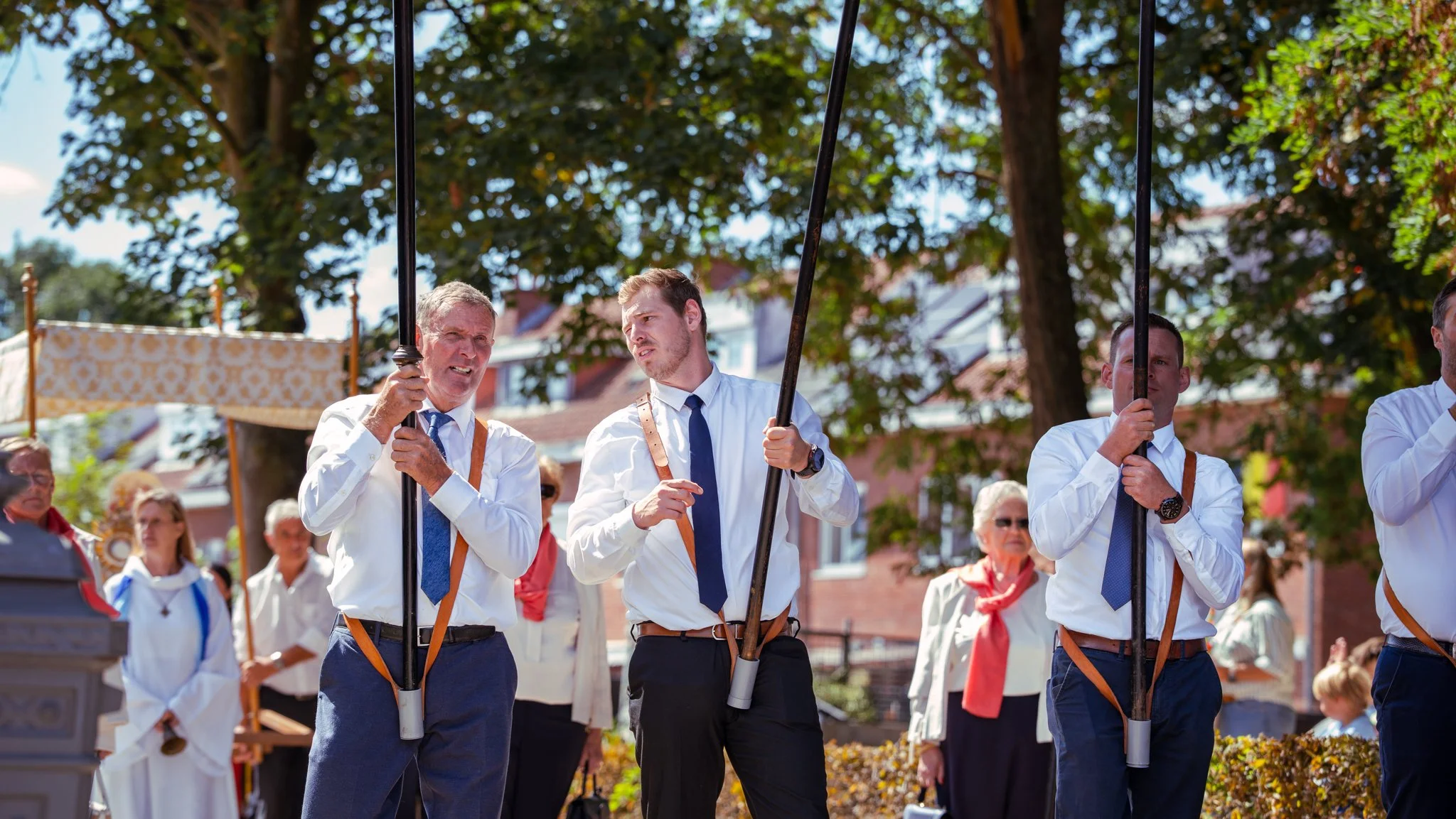 Group of men in white shirts and blue ties, holding long poles, participating in a parade or outdoor event on a sunny day, with trees and buildings in the background.