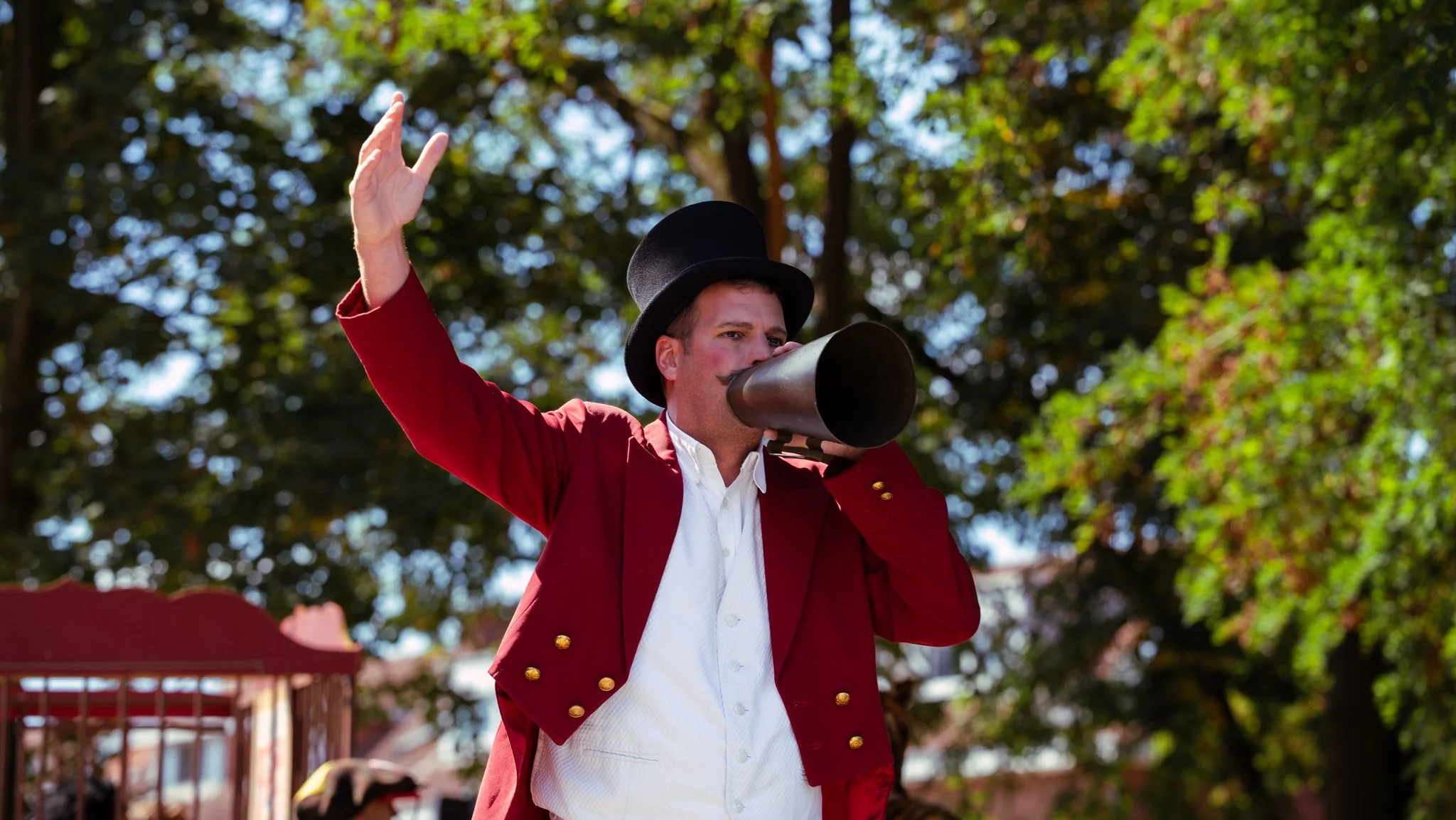 A man dressed as a ringmaster in a red jacket, white shirt, and black top hat, speaking into a megaphone outdoors with trees in the background.