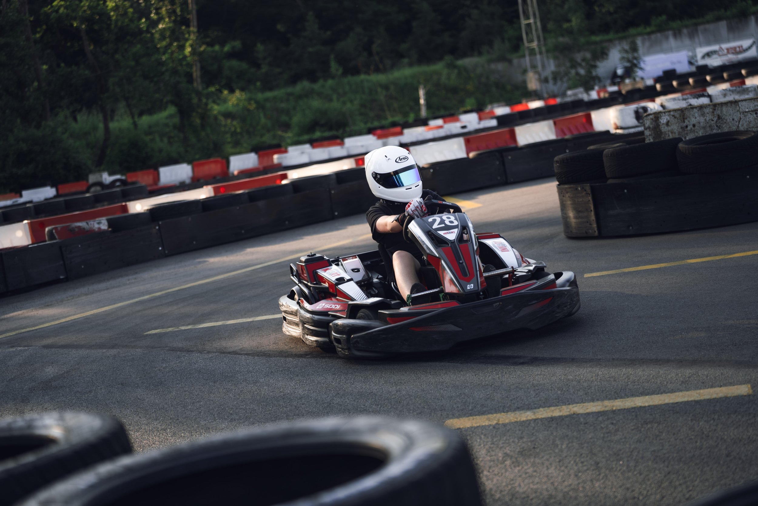A person racing go-karts on an outdoor track, wearing a white helmet, black shirt, and gray gloves, with barriers made of tires along the sides of the track.