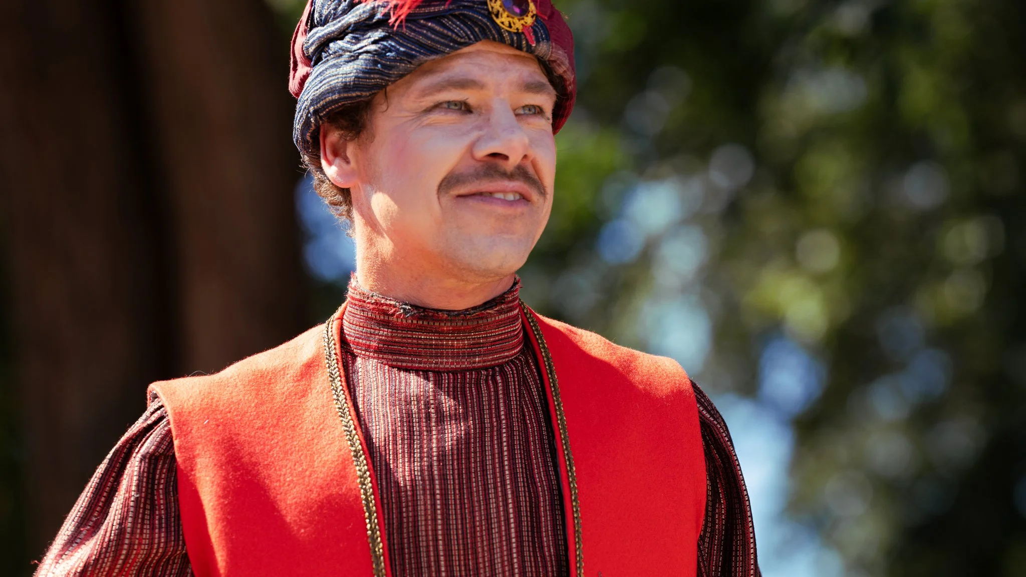 A man dressed in traditional clothing with a patterned hat, a red vest, and a striped shirt, standing outdoors with blurred trees in the background.