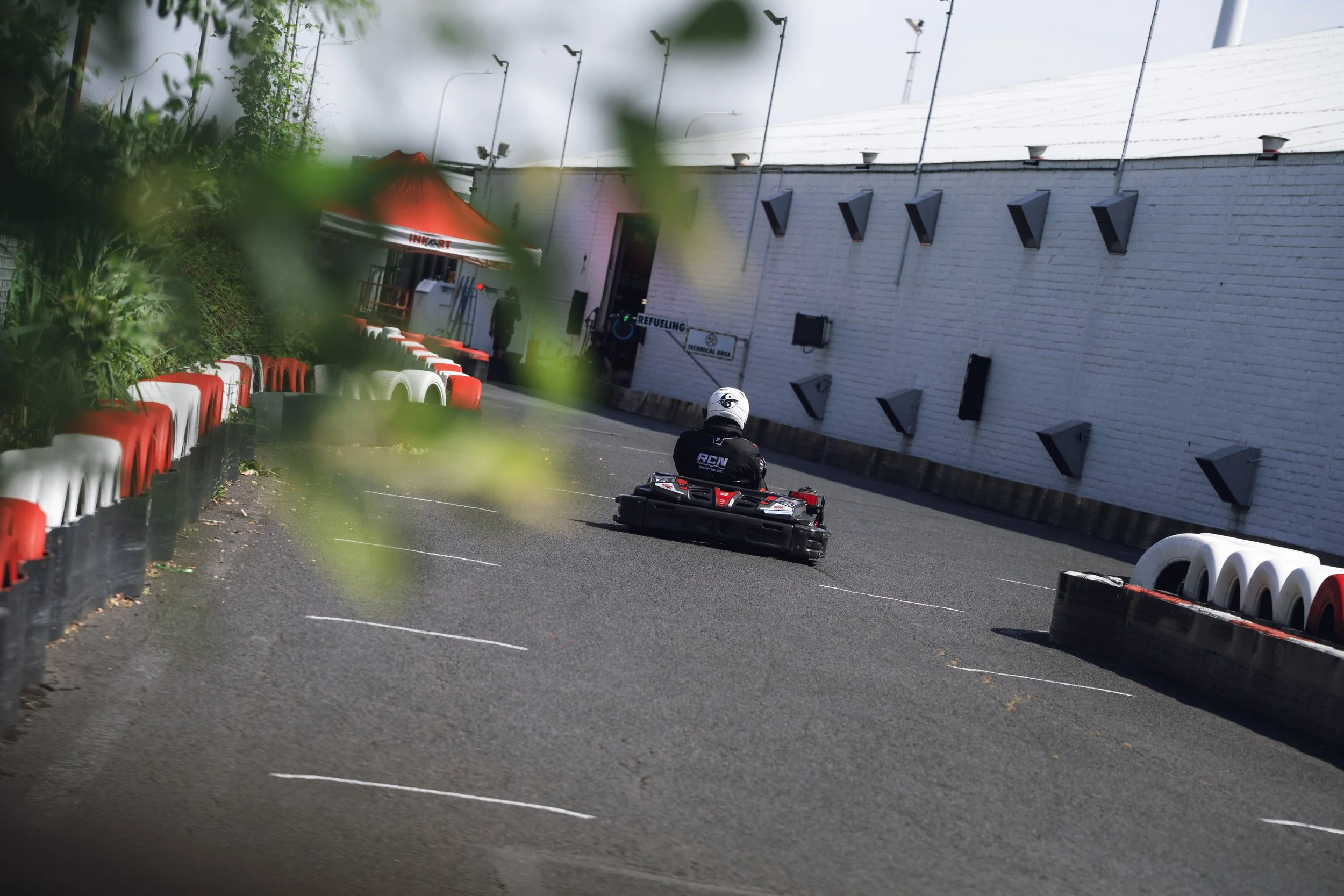 A person in a go-kart racing suit and helmet driving on a street circuit with barriers and a white wall with black vents, some greenery in the foreground.
