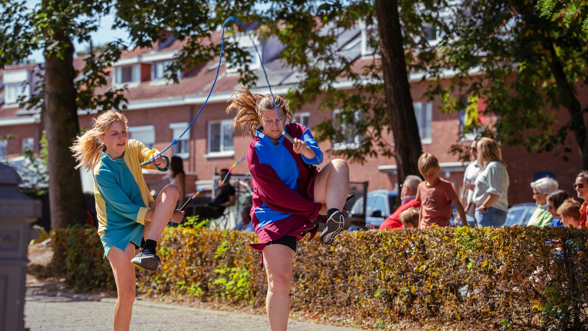 Two young girls jumping on a slackline outdoors during daytime with a group of people watching in the background.