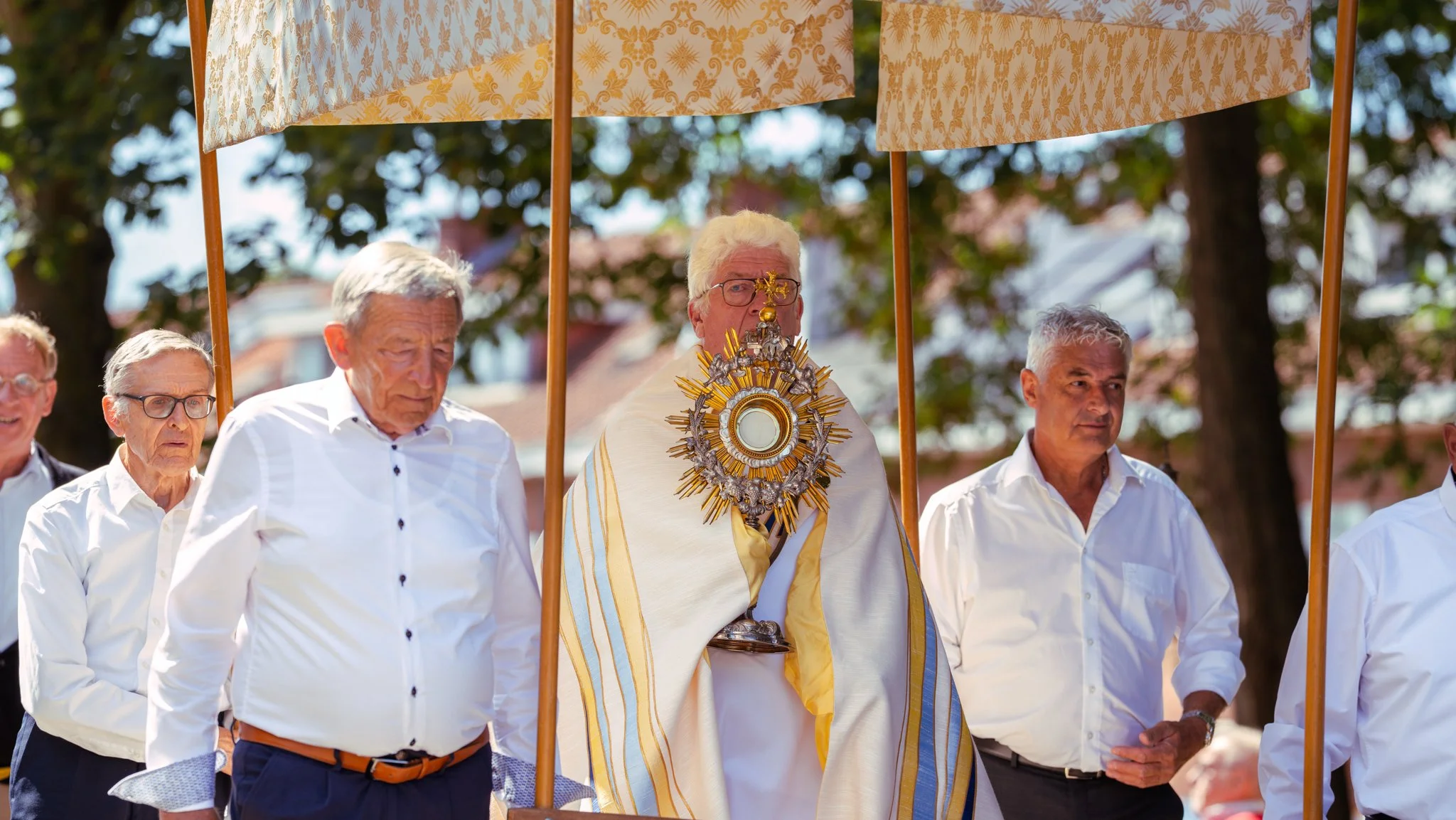 A religious procession with elderly men dressed in white shirts, carrying umbrellas, with a central figure wearing a cape adorned with a large ornate gold and silver religious relic or monstrance.