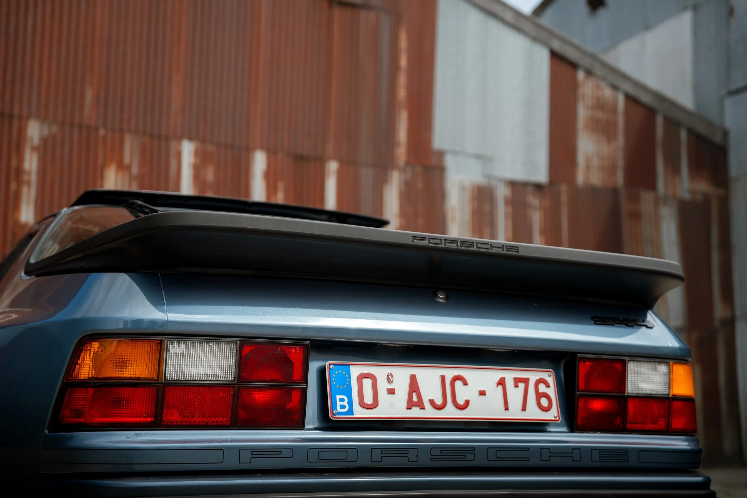 Rear view of a dark gray Porsche sports car with a roof rack, parked in front of a brick wall with a blue metal building in the background.