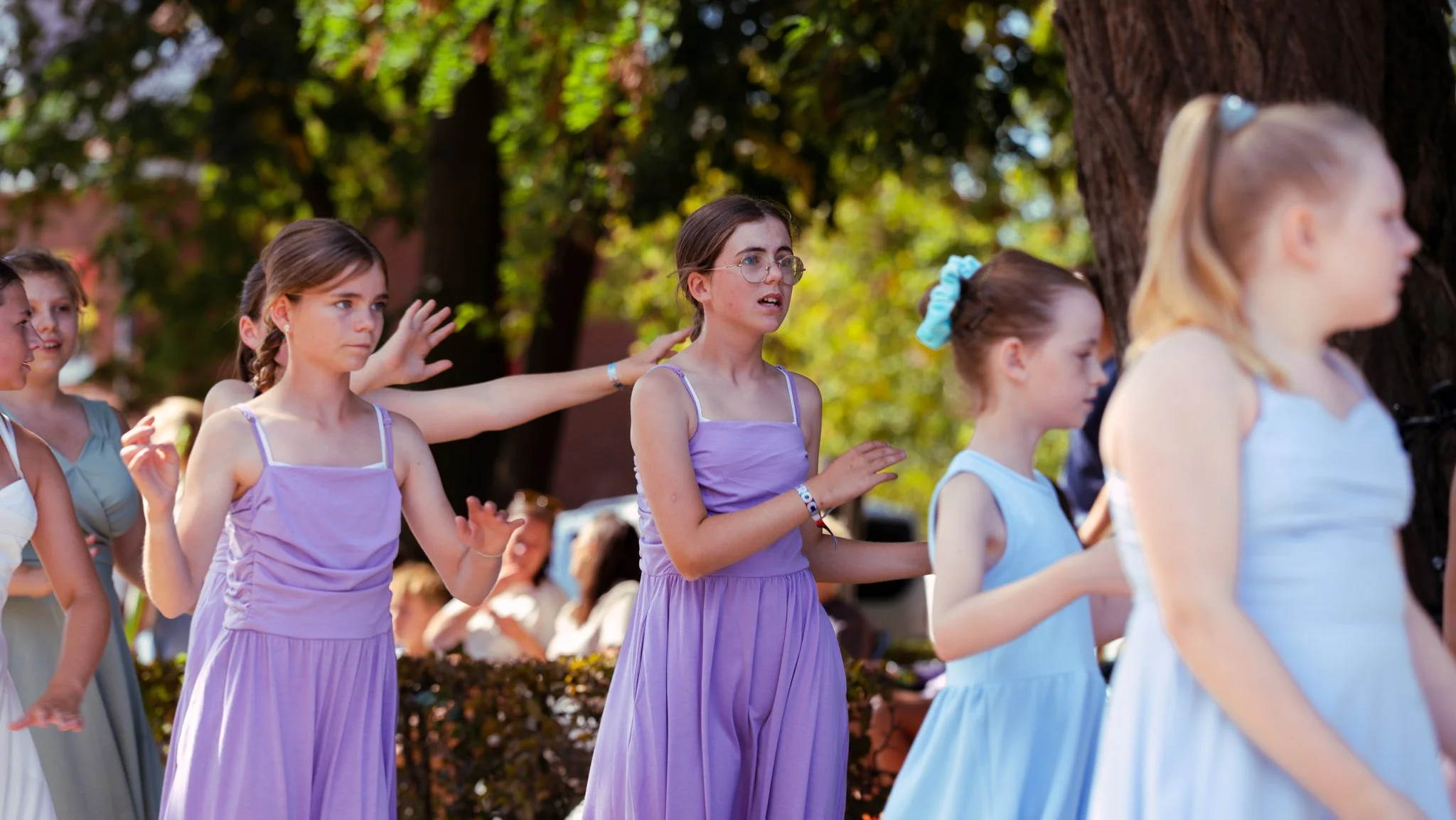 Group of young girls dressed in pastel-colored dresses standing outdoors under a large tree, some with arms outstretched and others with hands on their chests, participating in an activity or performance.