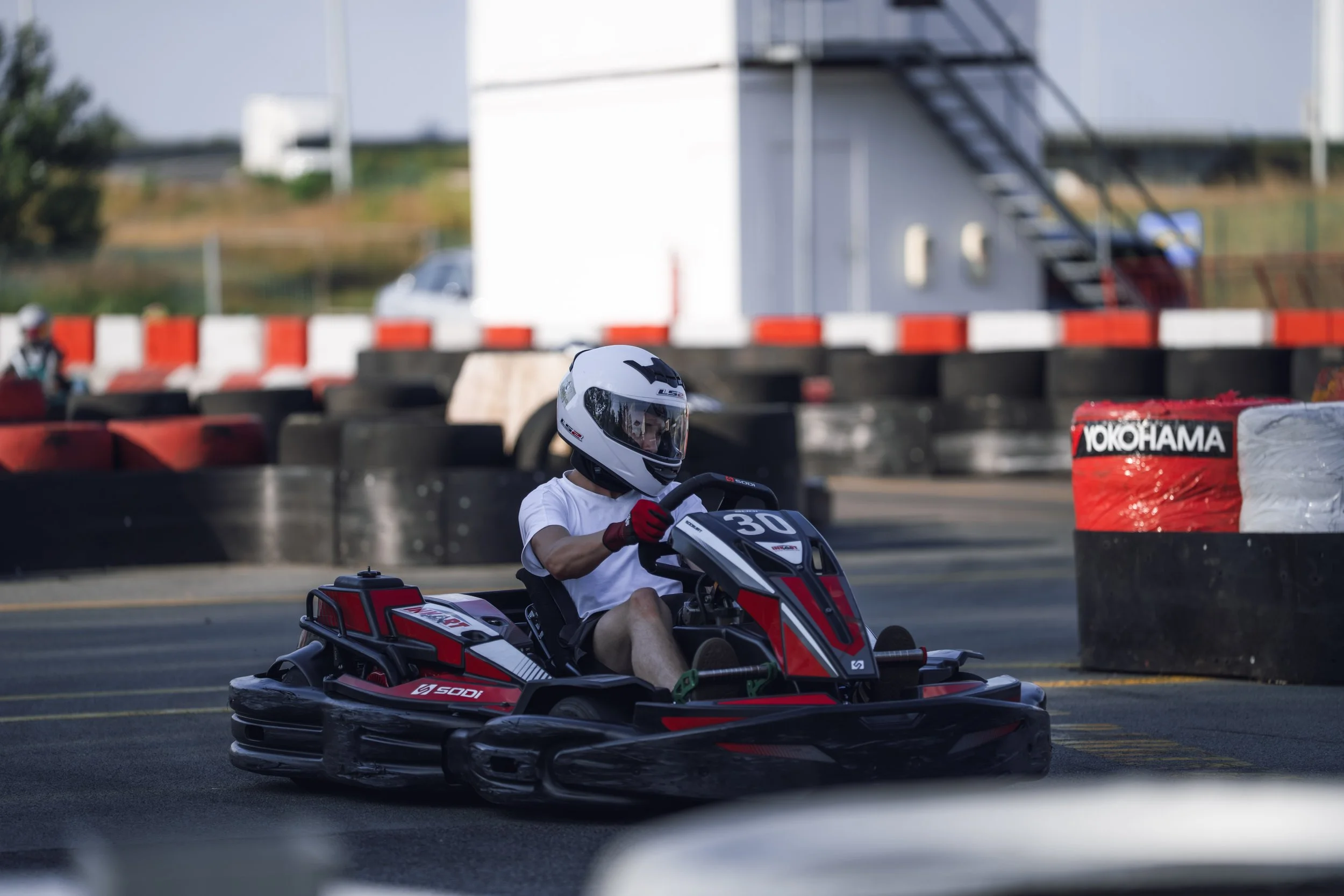 A person wearing a white helmet and red gloves riding a go-kart on an outdoor track with barriers and a building in the background.