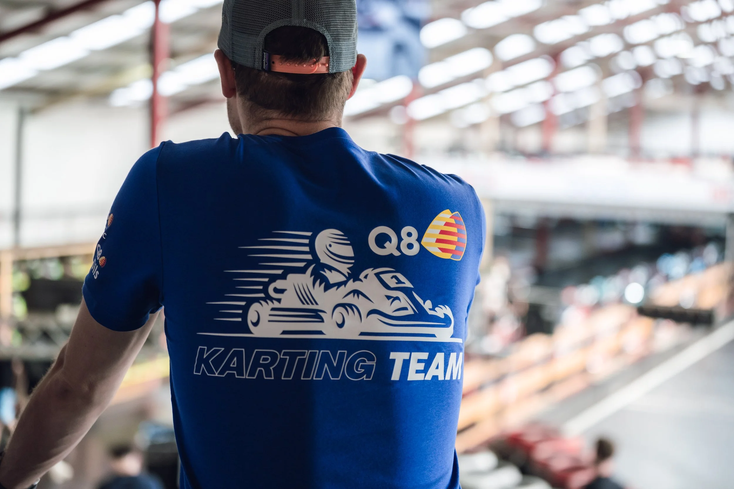 Person wearing a blue T-shirt with 'Karting Team' and a lion logo, facing away in a warehouse or garage filled with go-karts.