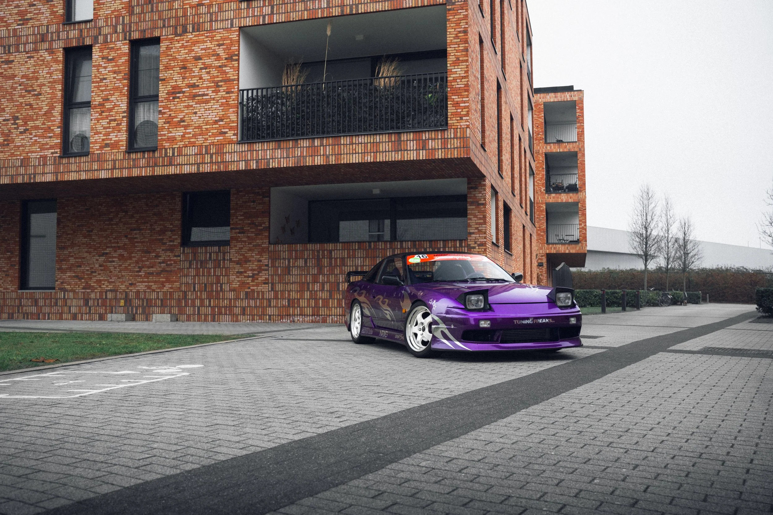 A purple sports car with customized decals is parked in front of a modern brick apartment building on a paved parking area.