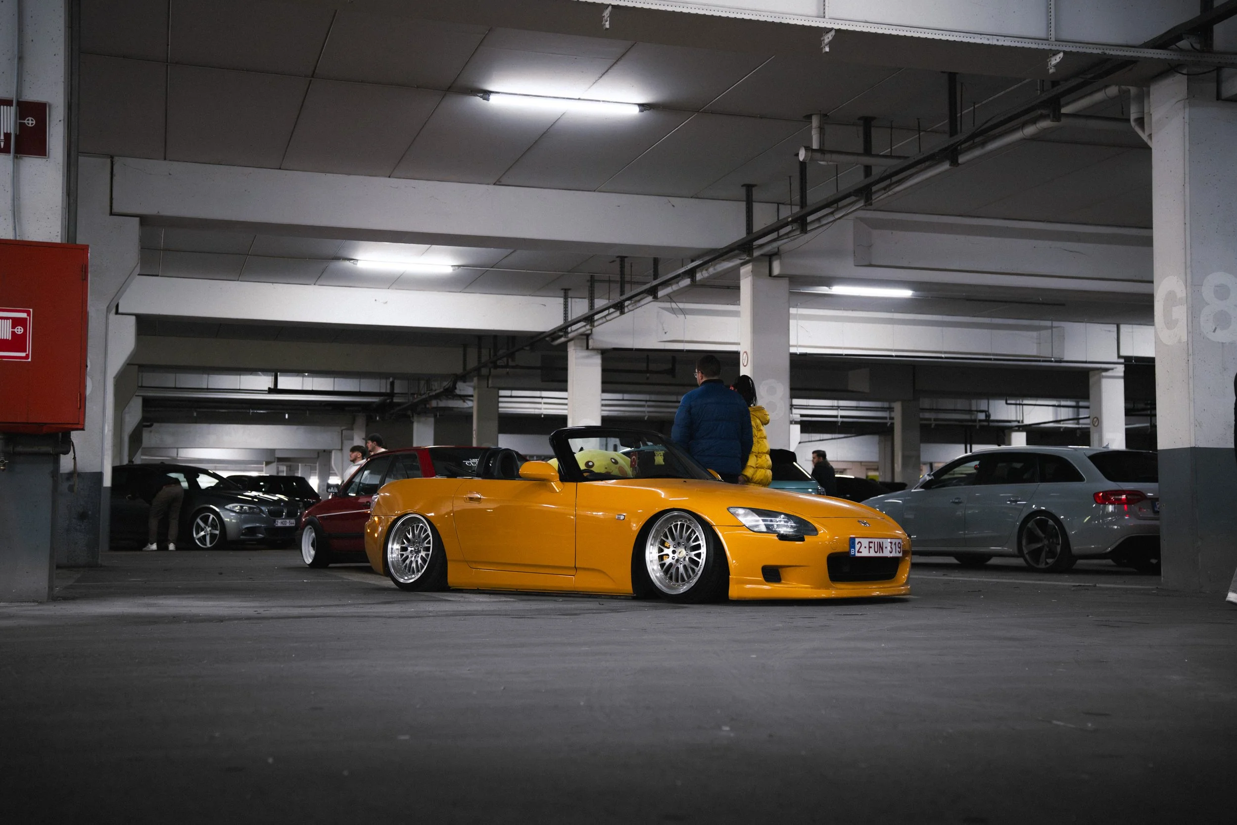 A bright yellow convertible sports car with custom wheels parked in an underground parking garage, with other cars and people in the background.