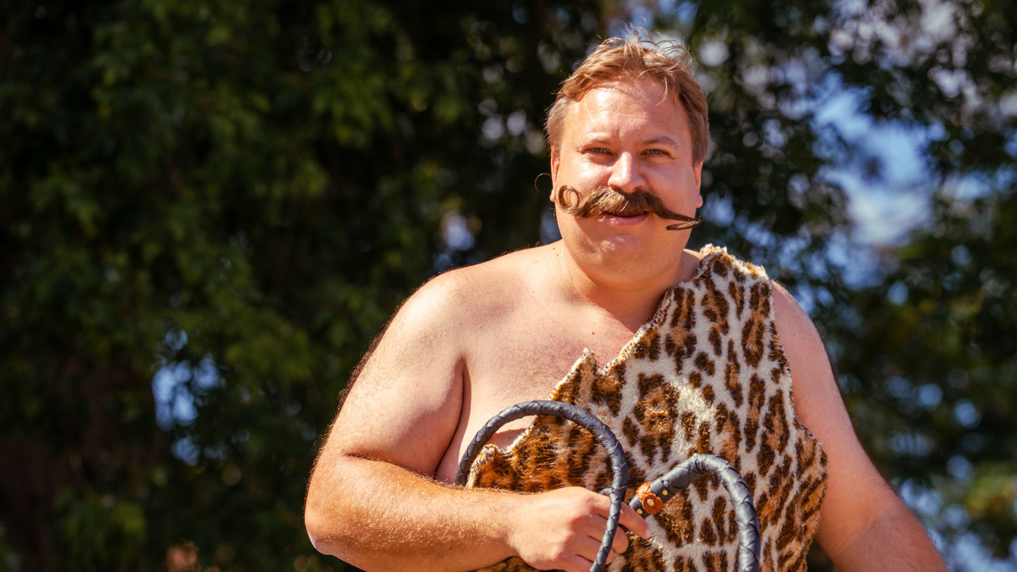 A man with a curly mustache and short hair smiling outdoors, wearing a leopard print fur cloak and holding a whip.
