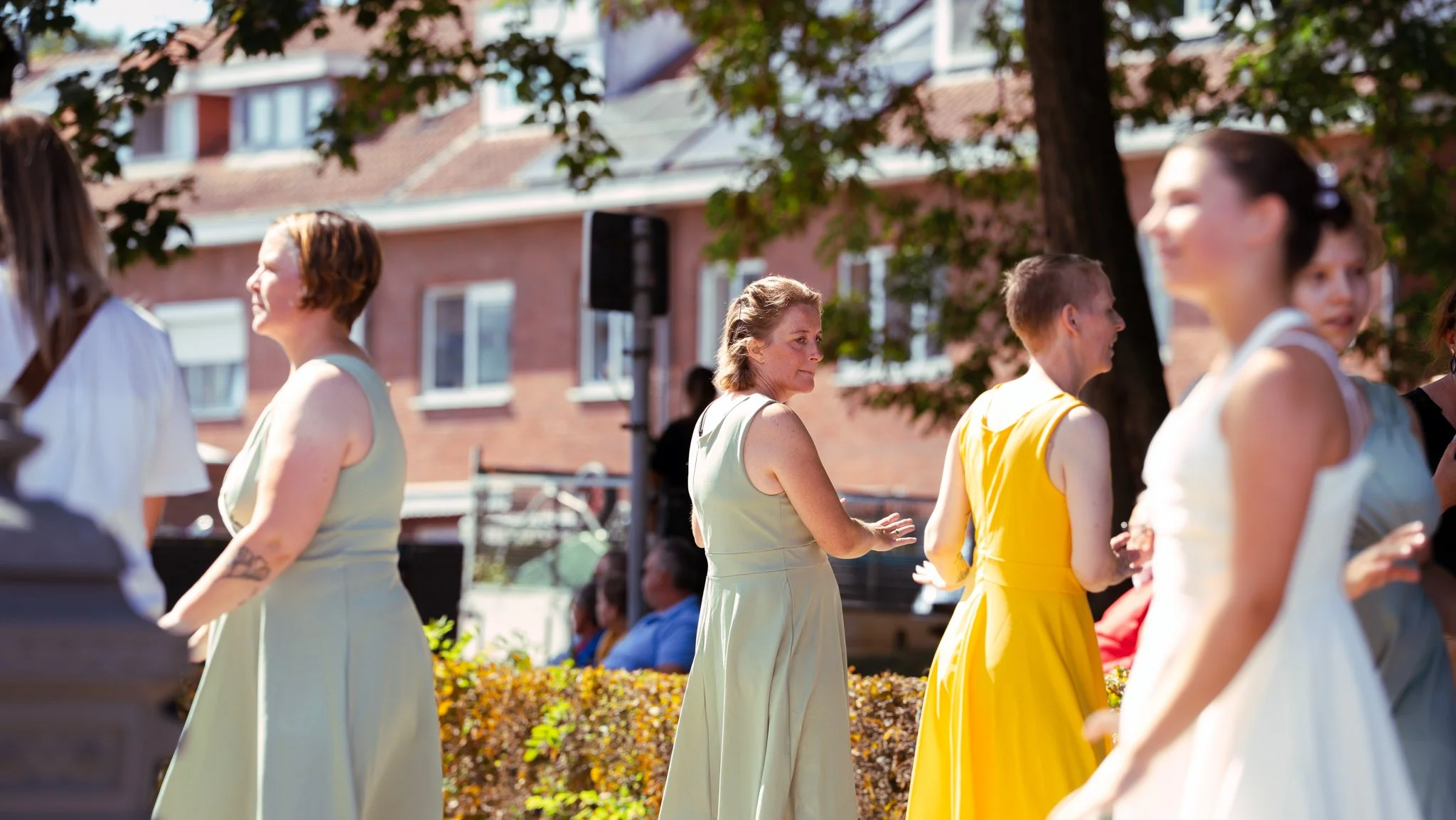People in pastel-colored dresses gathered outdoors on a sunny day, with a garden and brick buildings in the background.