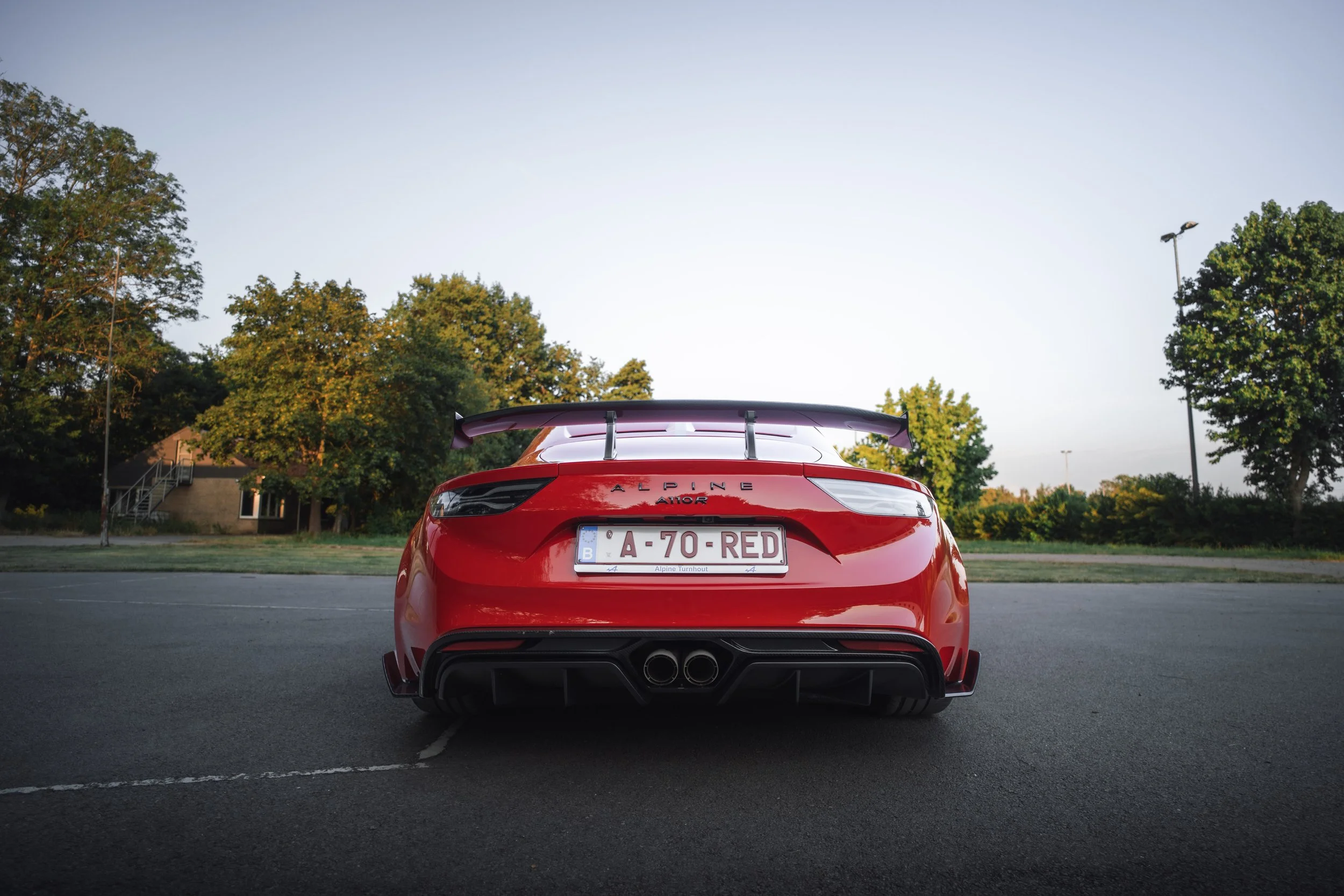 Rear view of a red Alpine A110 sports car parked in an empty parking lot with green trees and a building in the background, during late afternoon or early evening.