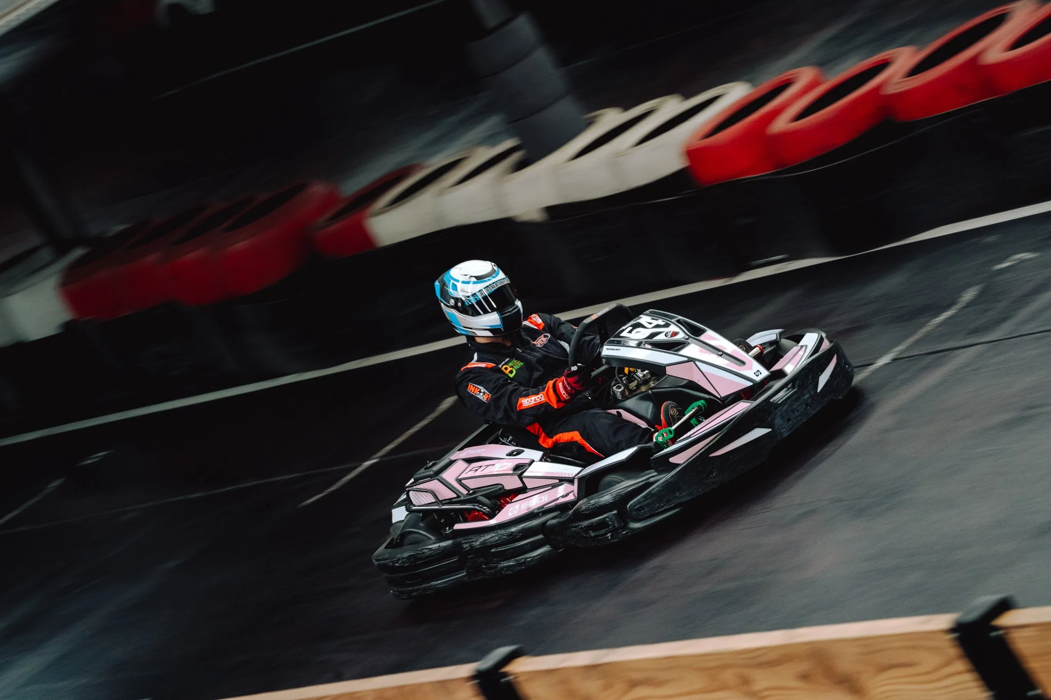 Person driving an electric go-kart on an indoor racing track, wearing a black racing suit with red accents and a blue helmet.