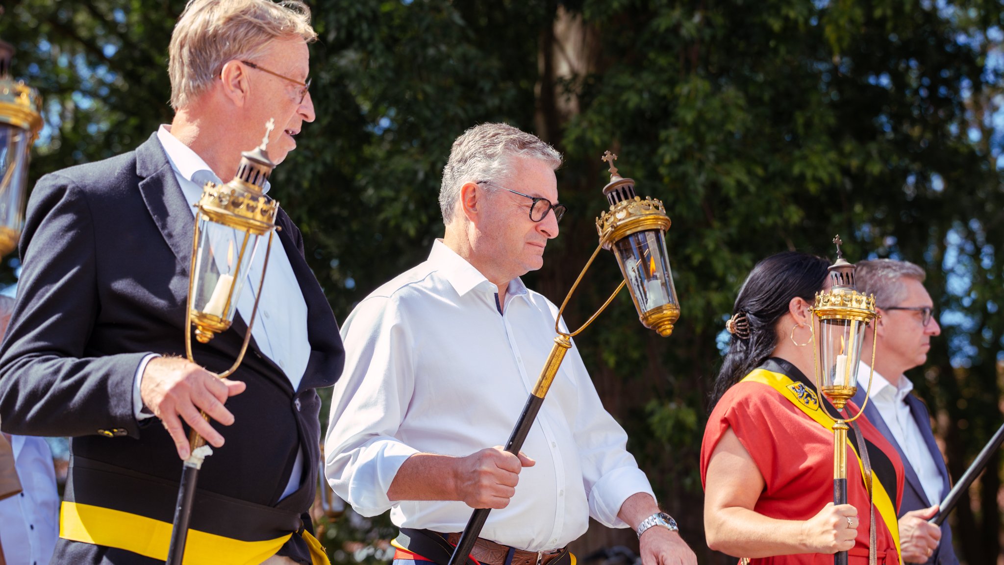 Group of people participating in a religious or memorial ceremony outdoors, holding lanterns with candles, standing in front of trees.