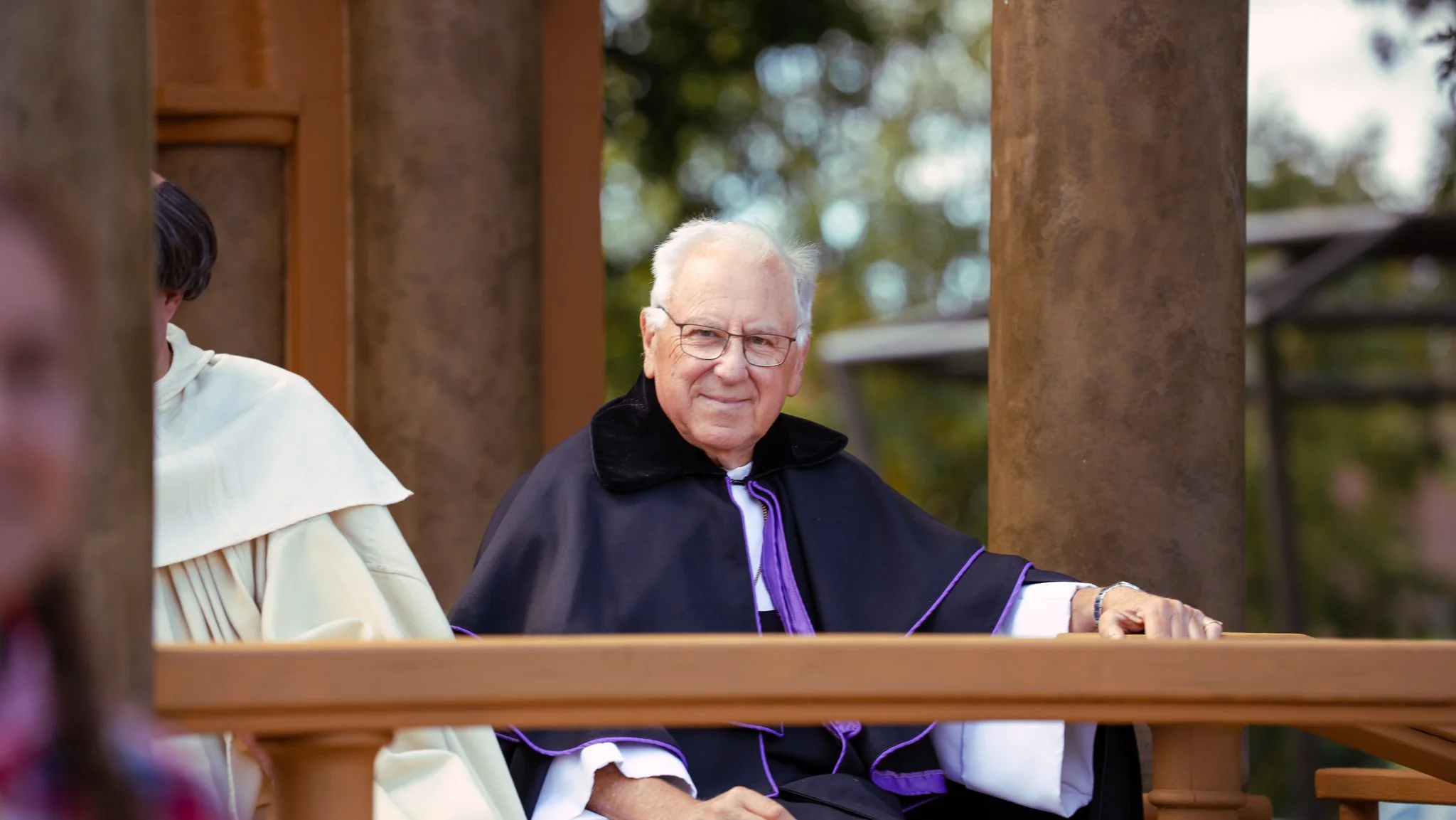 An elderly man with white hair and glasses, wearing a black graduation gown with purple accents, sitting outdoors on a wooden bench, surrounded by wooden pillars with blurred green trees in the background.