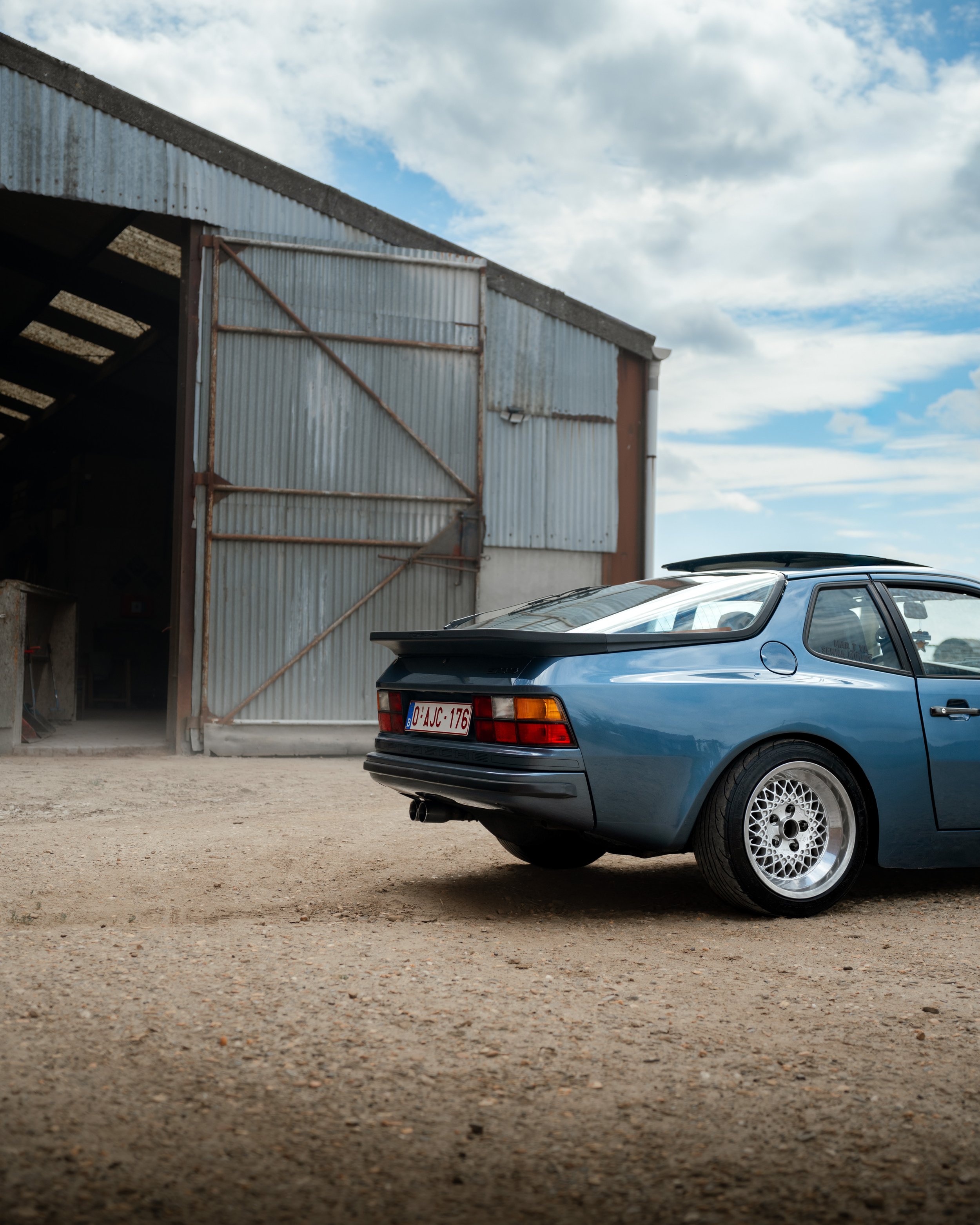 A blue sports car parked outside a rusted industrial building with a corrugated metal exterior, under a partly cloudy sky.