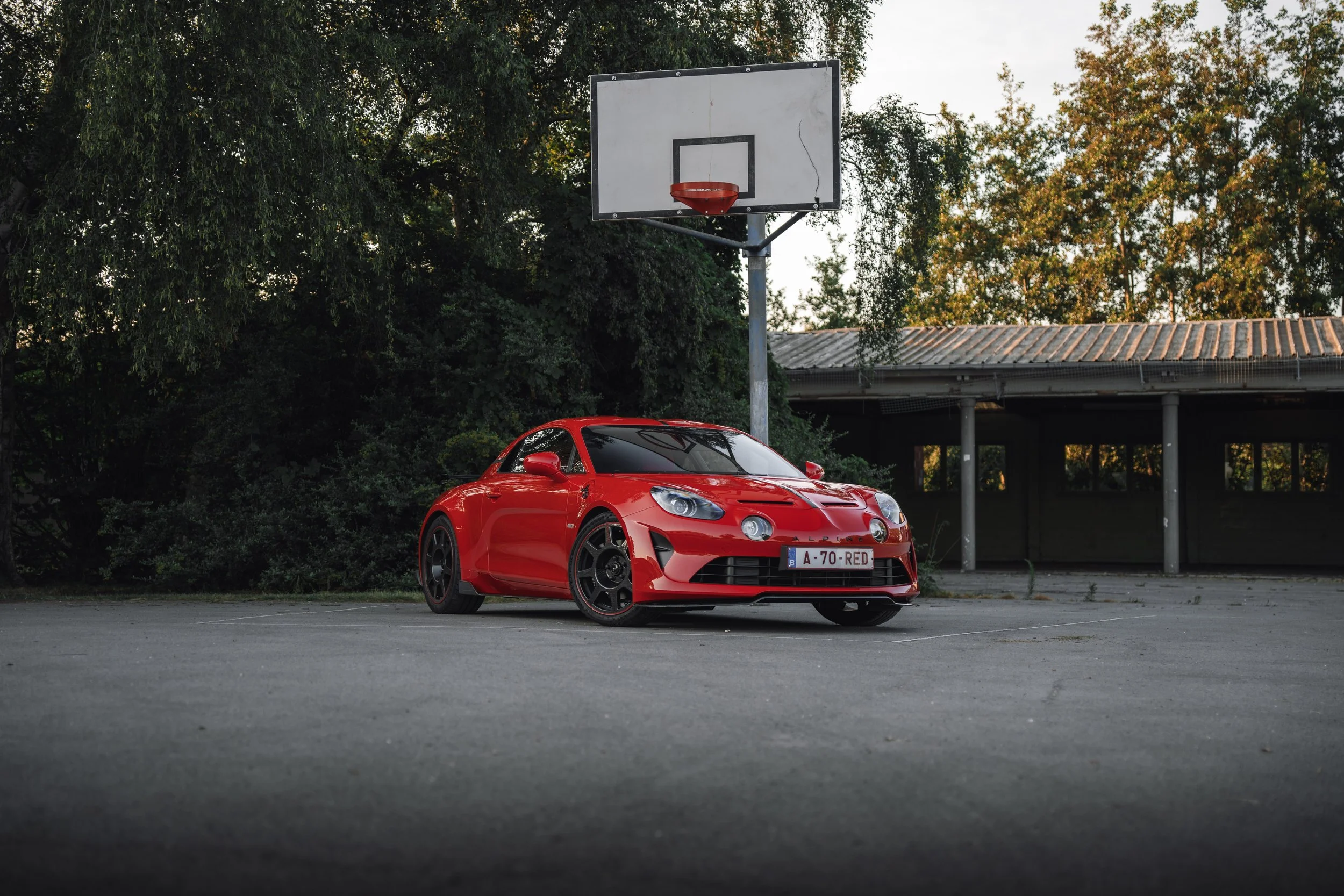A bright red sports car parked on a paved area near a basketball hoop with a white backboard and red rim. Trees with green and autumn-colored leaves are in the background.