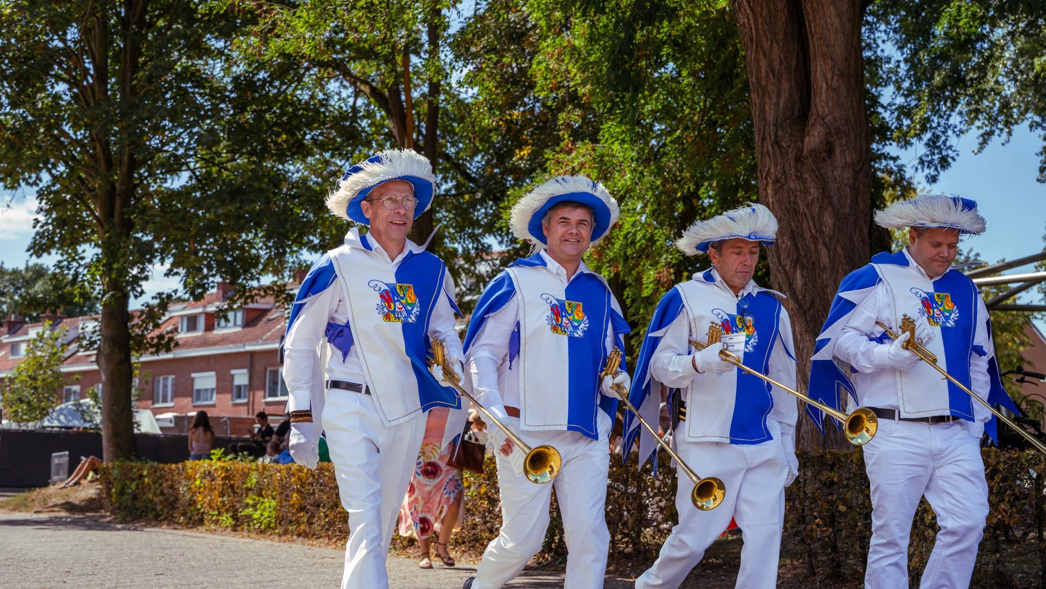 Four men dressed in blue and white uniforms with feathered hats, carrying brass instruments, walking outdoors on a sunny day.