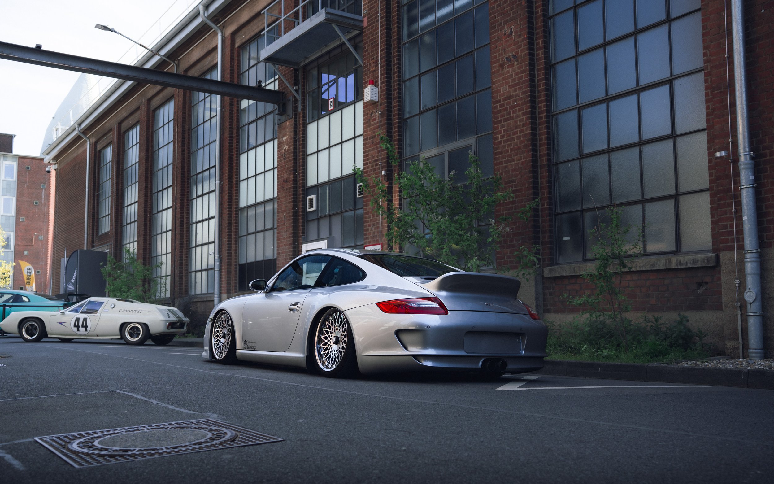 Several cars parked along a city street in front of a brick building with large industrial-style windows.